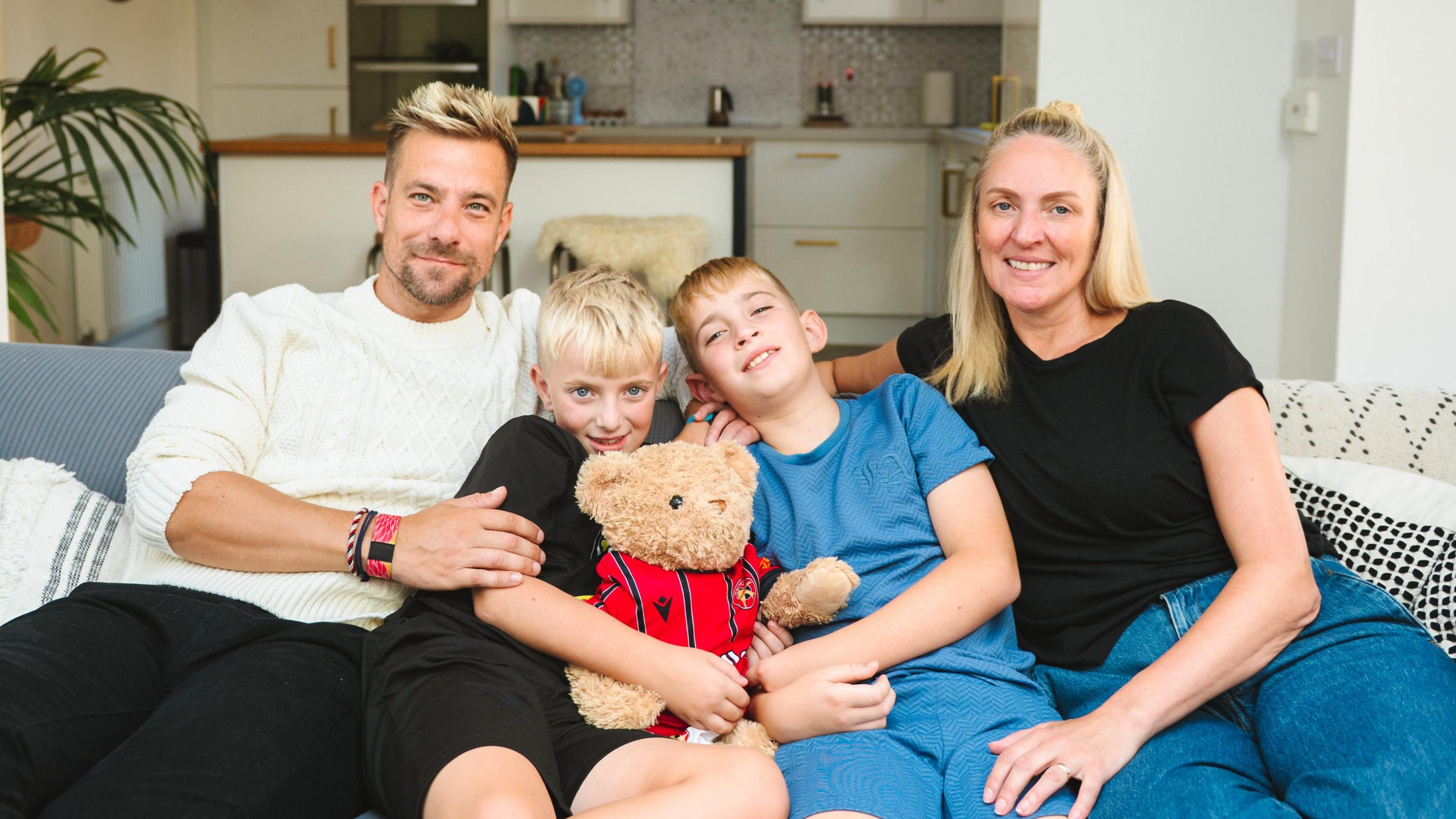 An image of a family, a mother and father and two young boys, sitting on a sofa in their home. In the background you can see their kitchen. In between the young boys there is a brown teddy bear wearing a red football top