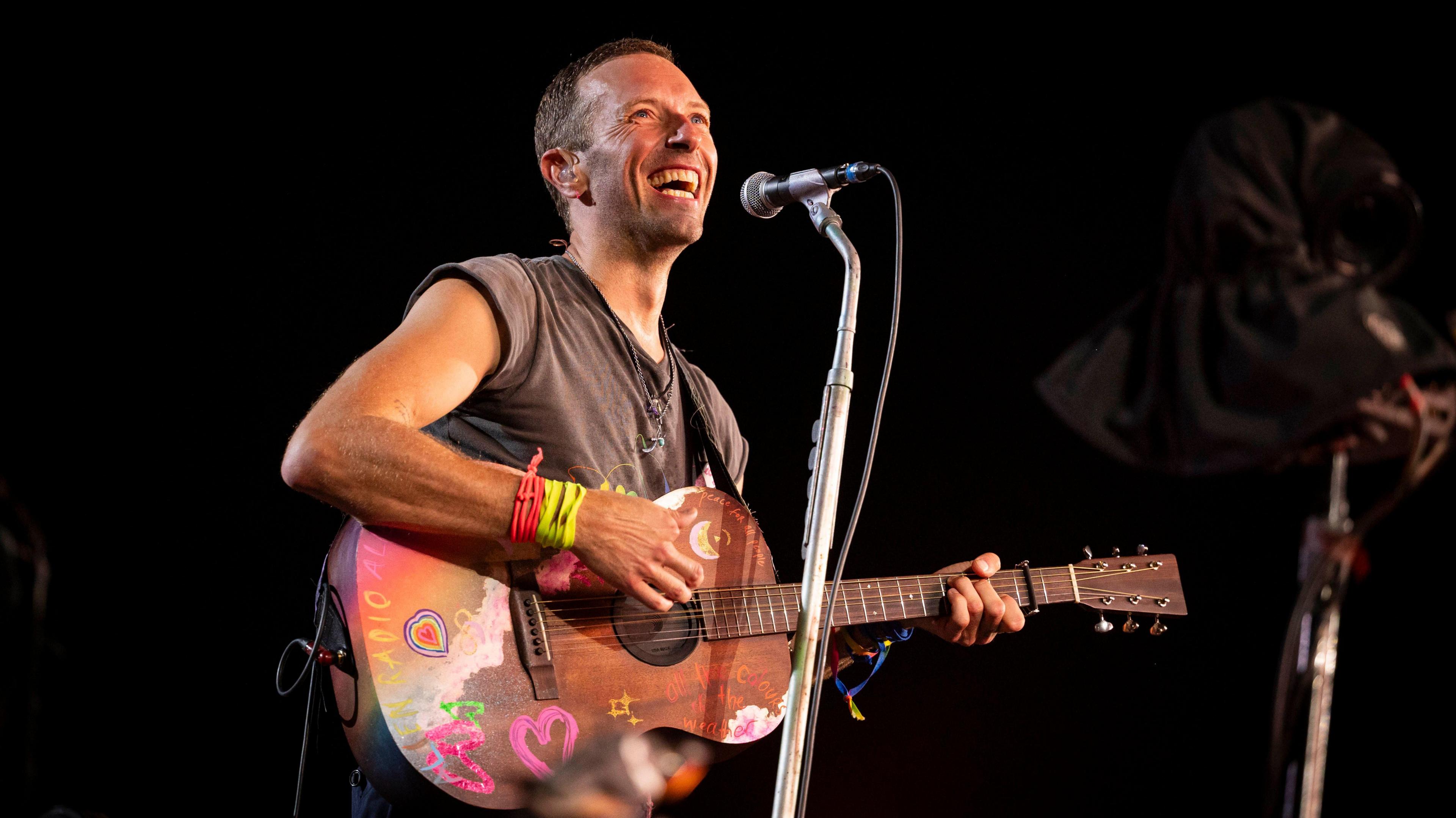 A man - Chris Martin - smiling wearing a grey t-shirt and holding a wooden guitar with colourful doodles on. A microphone is on a stand in front of him and the background is dark.