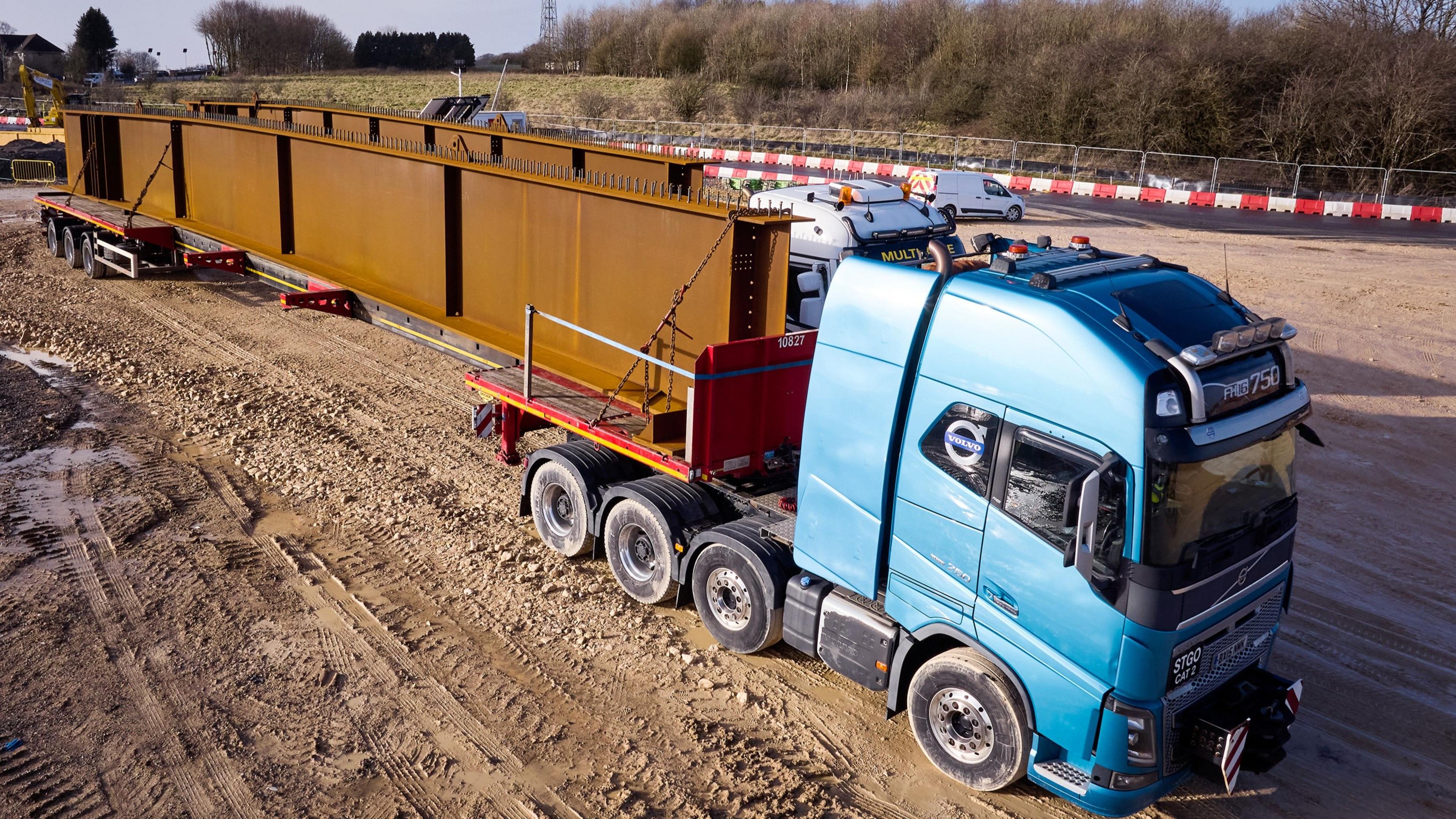 A blue lorry driving through a muddy section of a construction site. On its bed, which has been split into two, there is a large copper-coloured beam. 