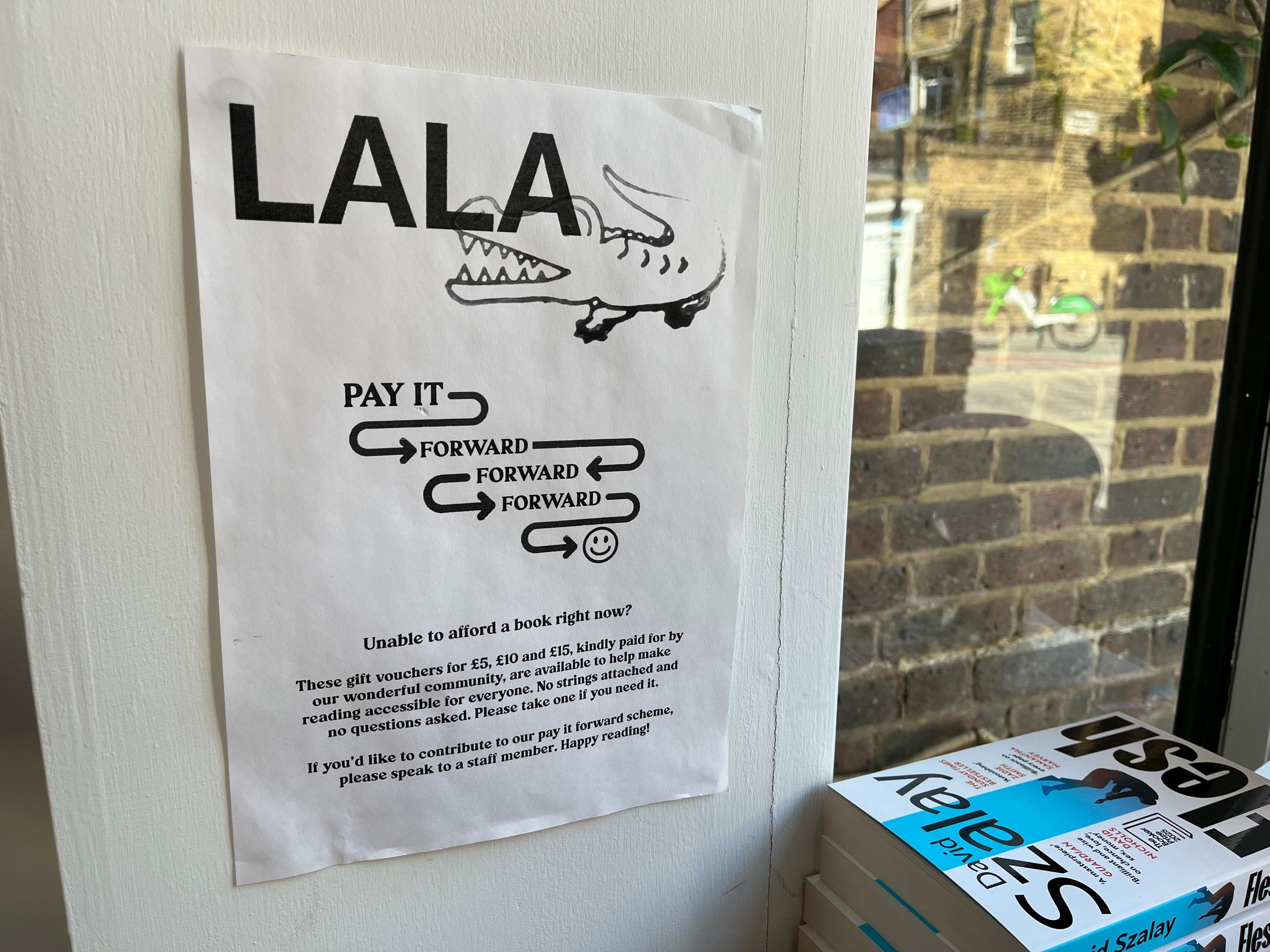 A sign with the name LALA and an image of a crocodile. Text below reads: Pay it forward, forward, forward with arrows between the words and a smiley face. The sign also says: Unable to afford a book right now? These gift vouchers for £5, £10, and £15, kindly paid for by our wonderful community are available to help make reading accessible for everyone. No strings attached and no questions asked. Please take one if you need it. If you'd like to contribute to our pay it forward scheme, please speak to a staff member. Happy reading! 