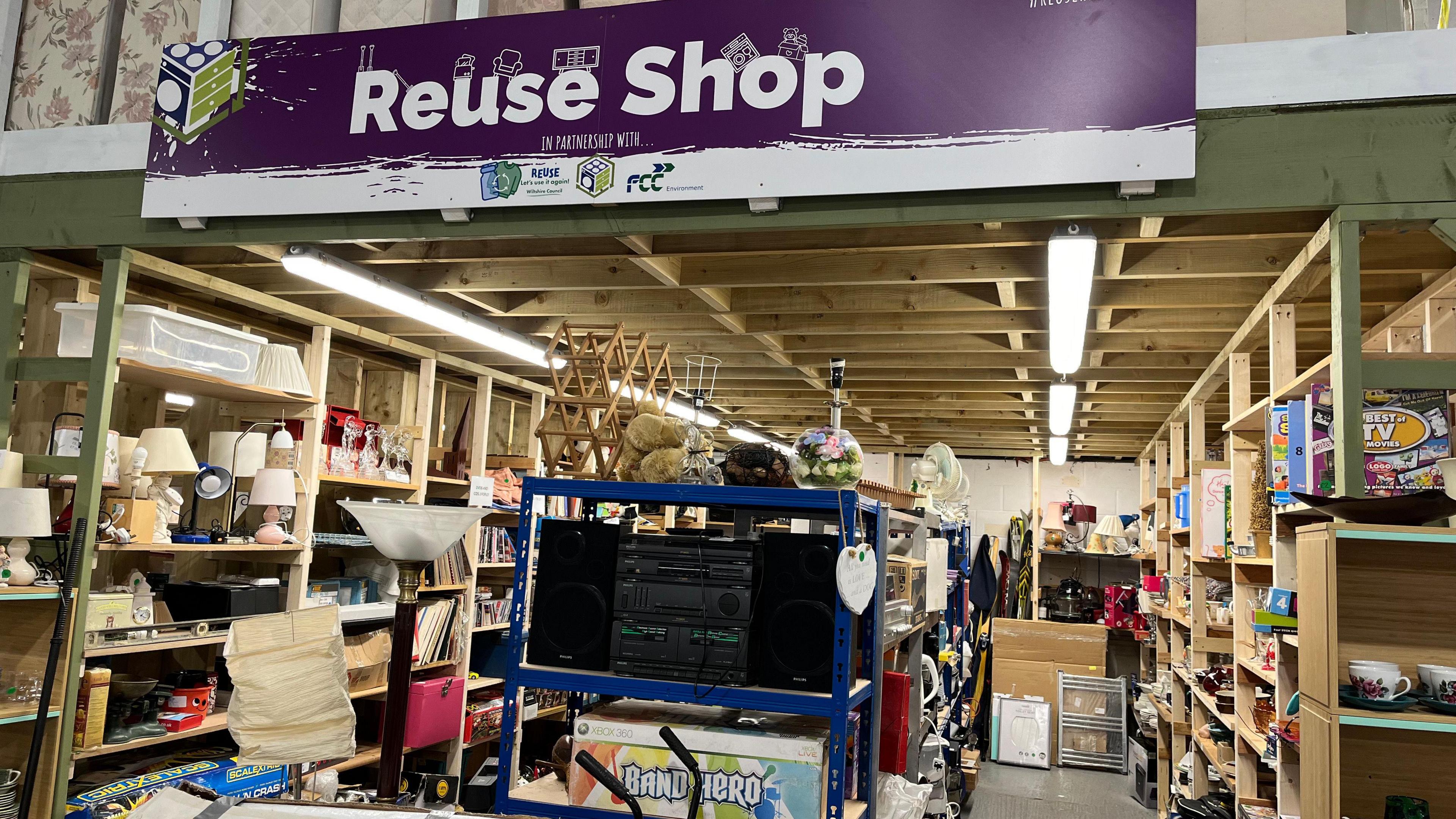 A view of the front of the shop- large sign above and rows of wooden shelves with a variety of items. Mostly bric-a-bric and small electricals.