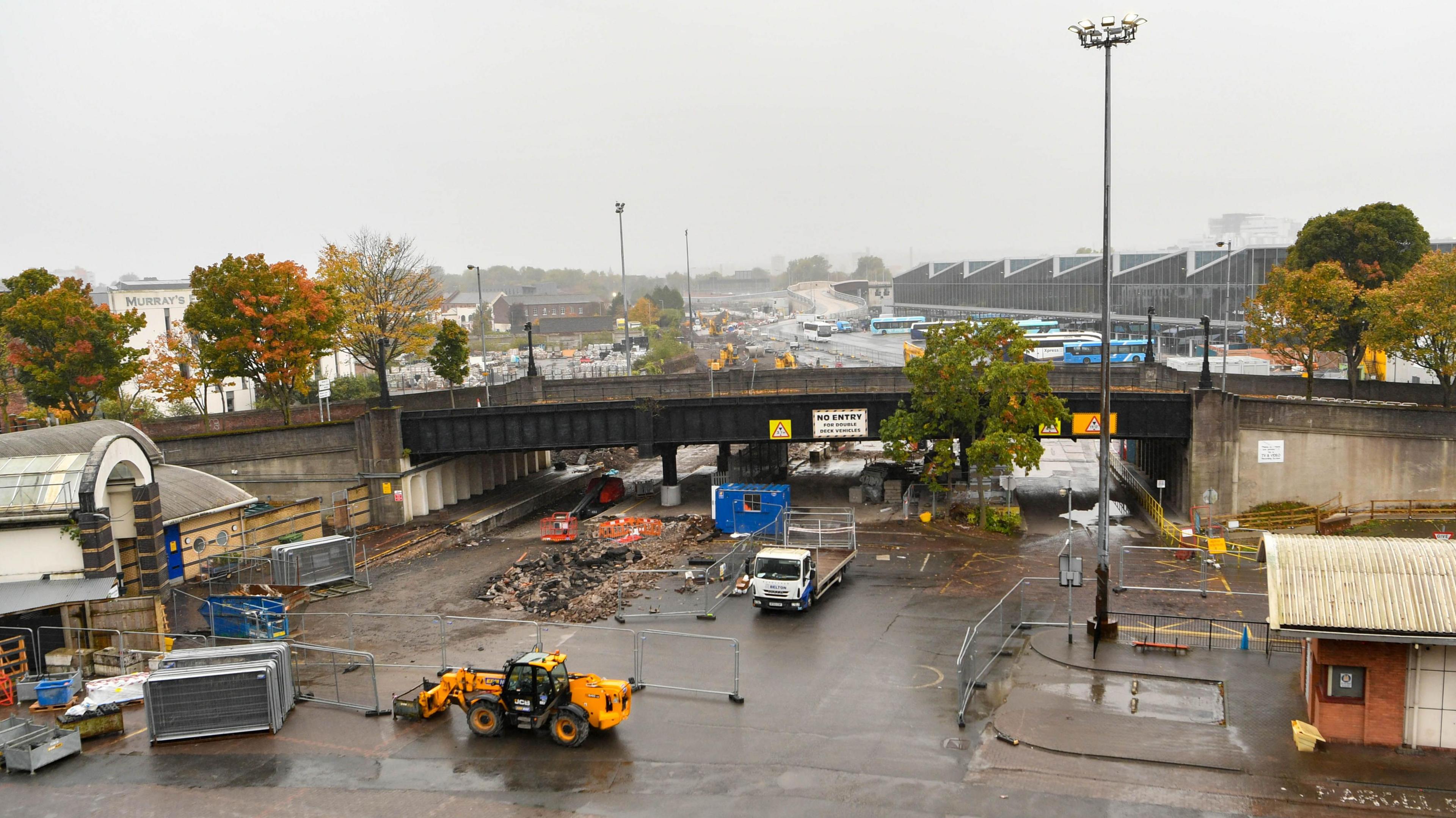 A side view of the Boyne Bridge in Belfast with the new Grand Central Station in the background.  The road under the bridge is partially cordoned off with steel barriers and there is a digger and a truck in the foreground. 