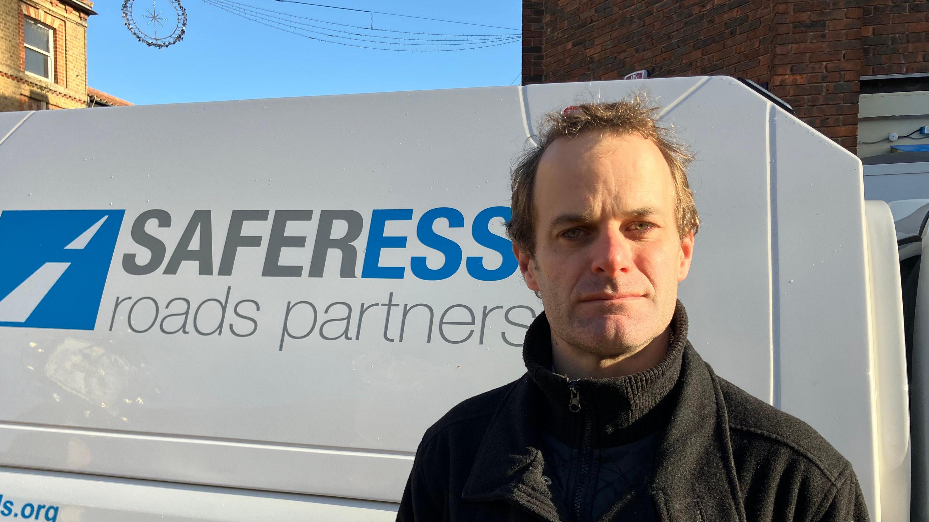 A man in front of a white van, wearing a black coat. The van is printed with the words "Safer Essex roads partnership".