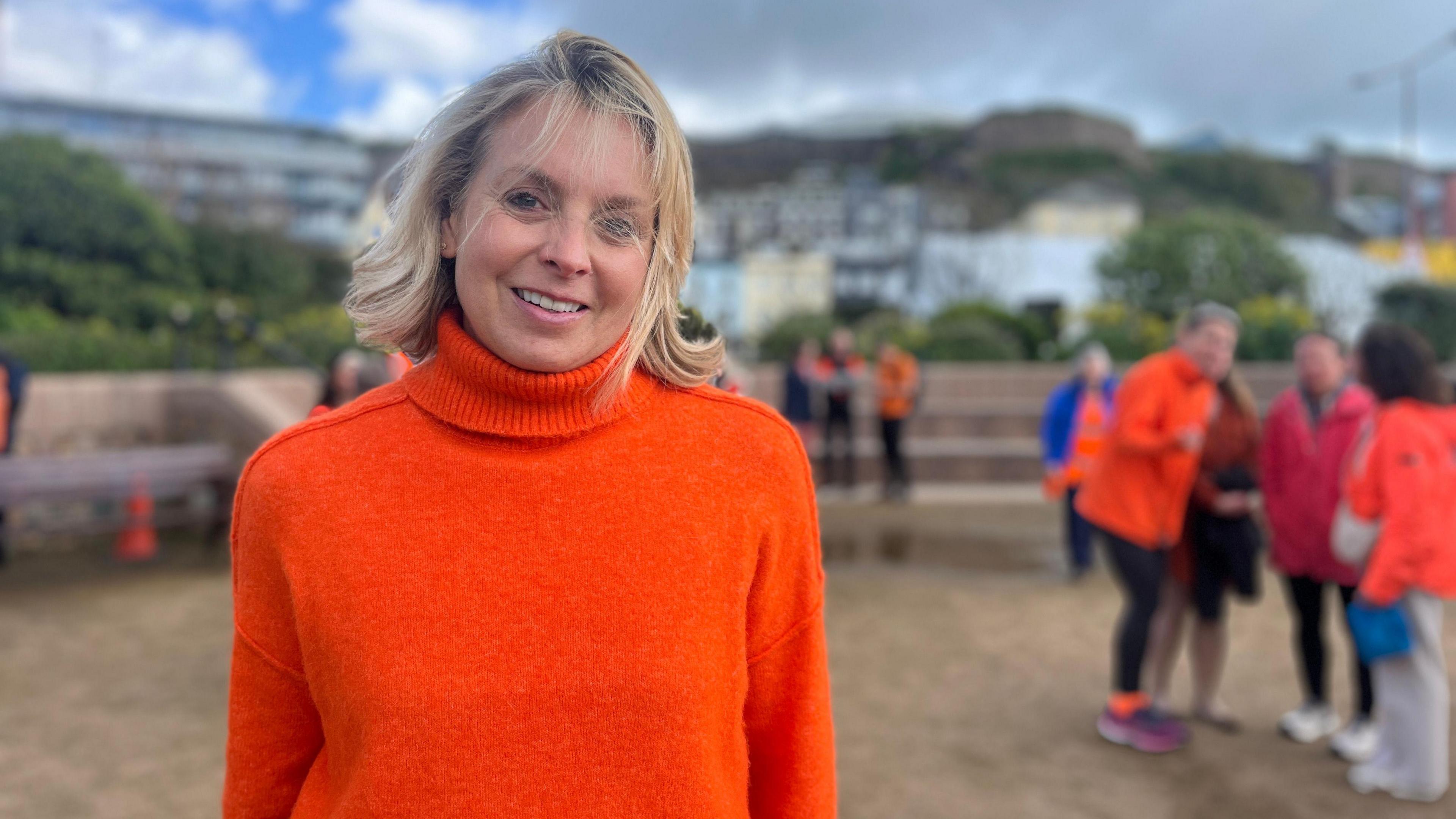 Claire smiles at the camera as people in orange chat behind her. She's wearing an orange turtleneck jumper. She has short blonde hair.