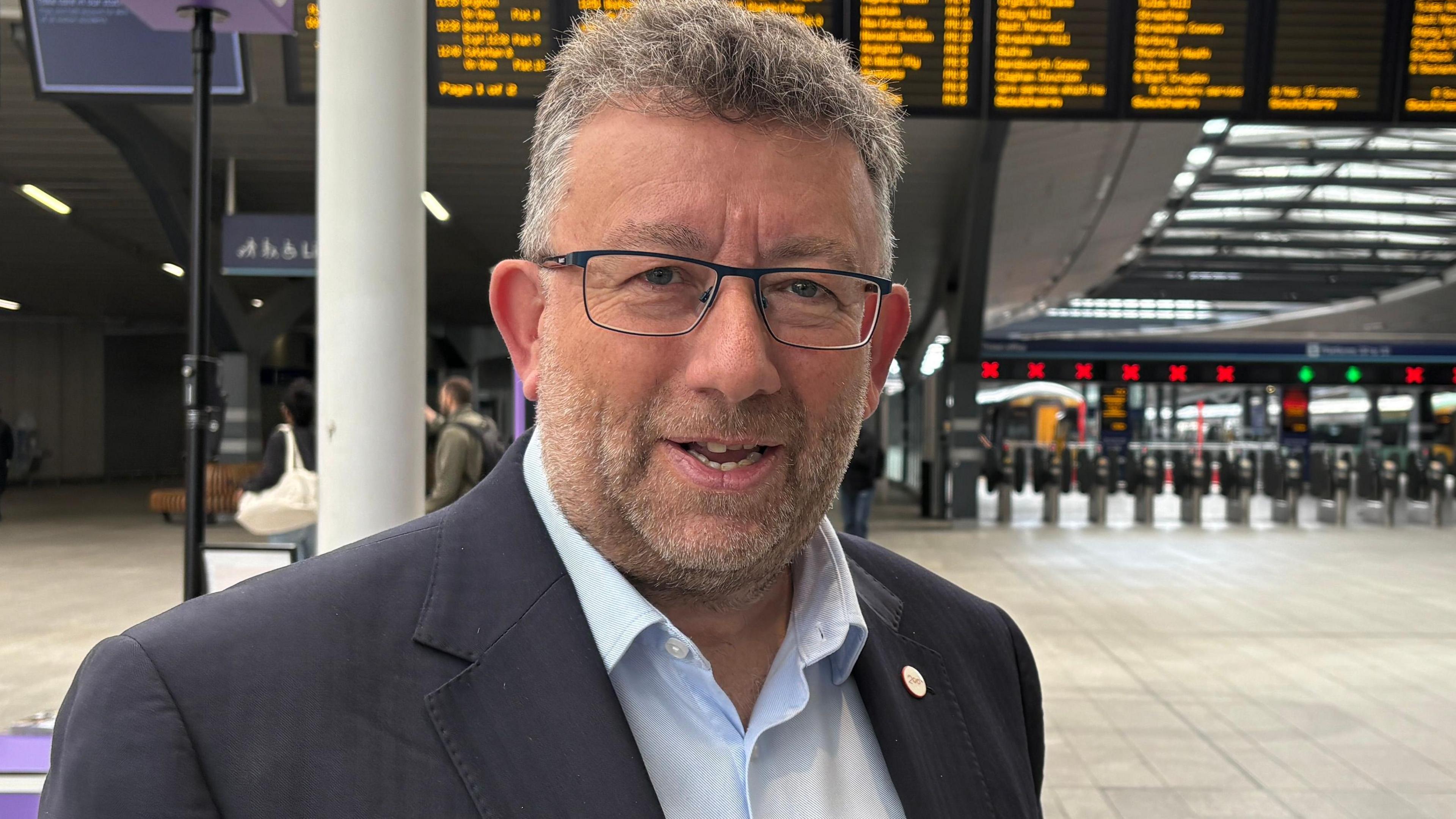Steve White looking directly down the camera. He is standing with his shoulders turned slightly away from the camera, but his head is turned towards it. He is wearing rectangular glasses, a light blue opened-necked shirt and a dark coloured blazer. He has a short, greying beard and short hair. He is standing inside a train station. There are information boards with orange writing on them suspended high up in the background. Beneath them is a row of turnstiles, many of which have red Xs over them; two have green ticks above them. There are a handful of passengers behind Mark, but otherwise, the station is reasonably quiet.