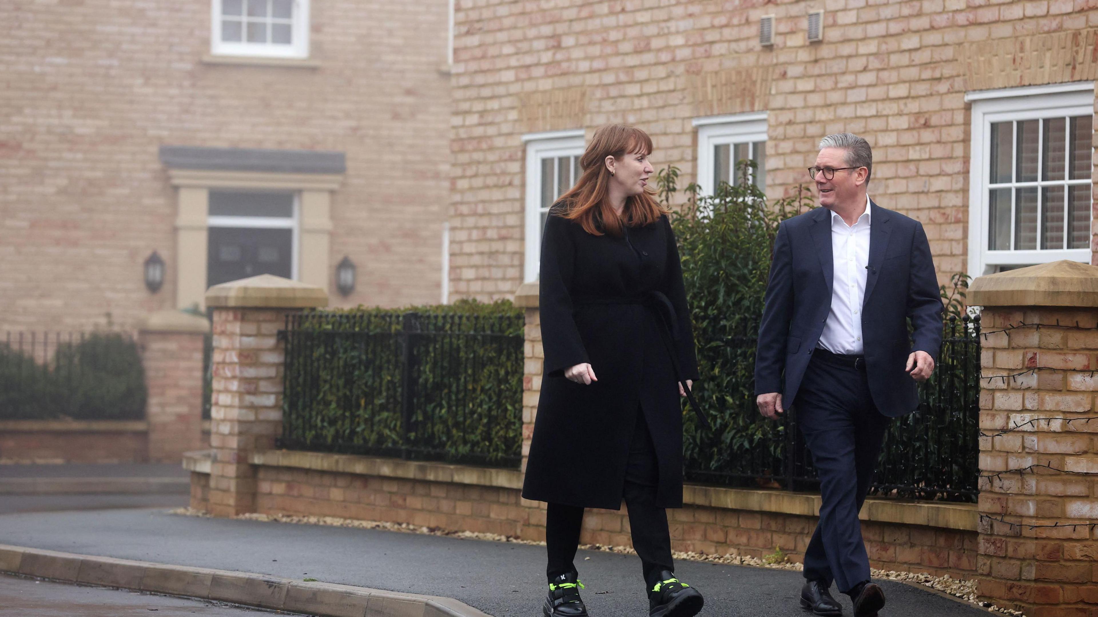 Angela Rayner and Sir Keir Starmer walk down a new build street. Angela wears a long black coat. Sir Keir wears a black suit with a white shirt with the top button undone. 