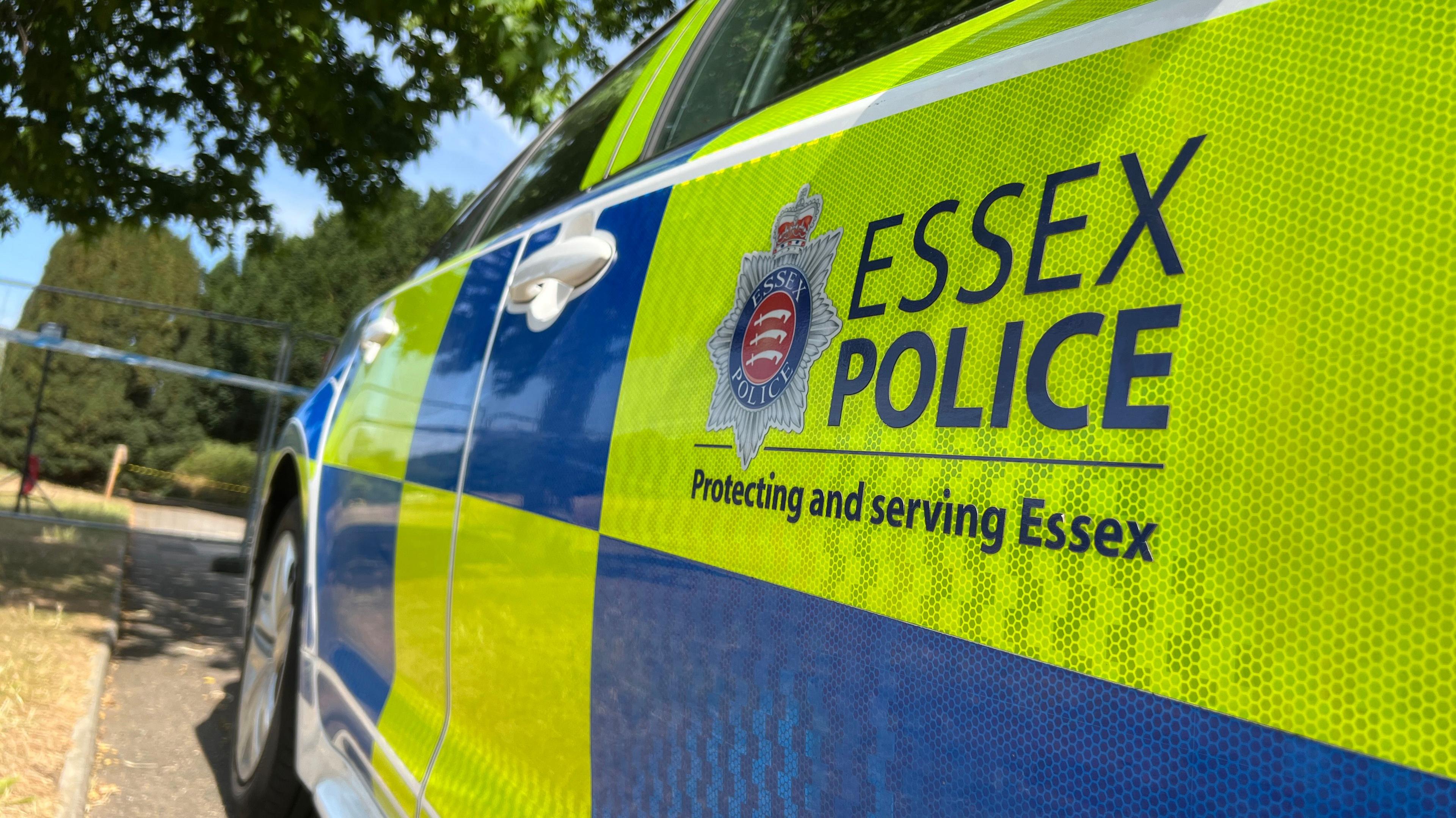 A low-angle shot of the side of an Essex Police car parked by a police cordon. The car has yellow and blue squares and the