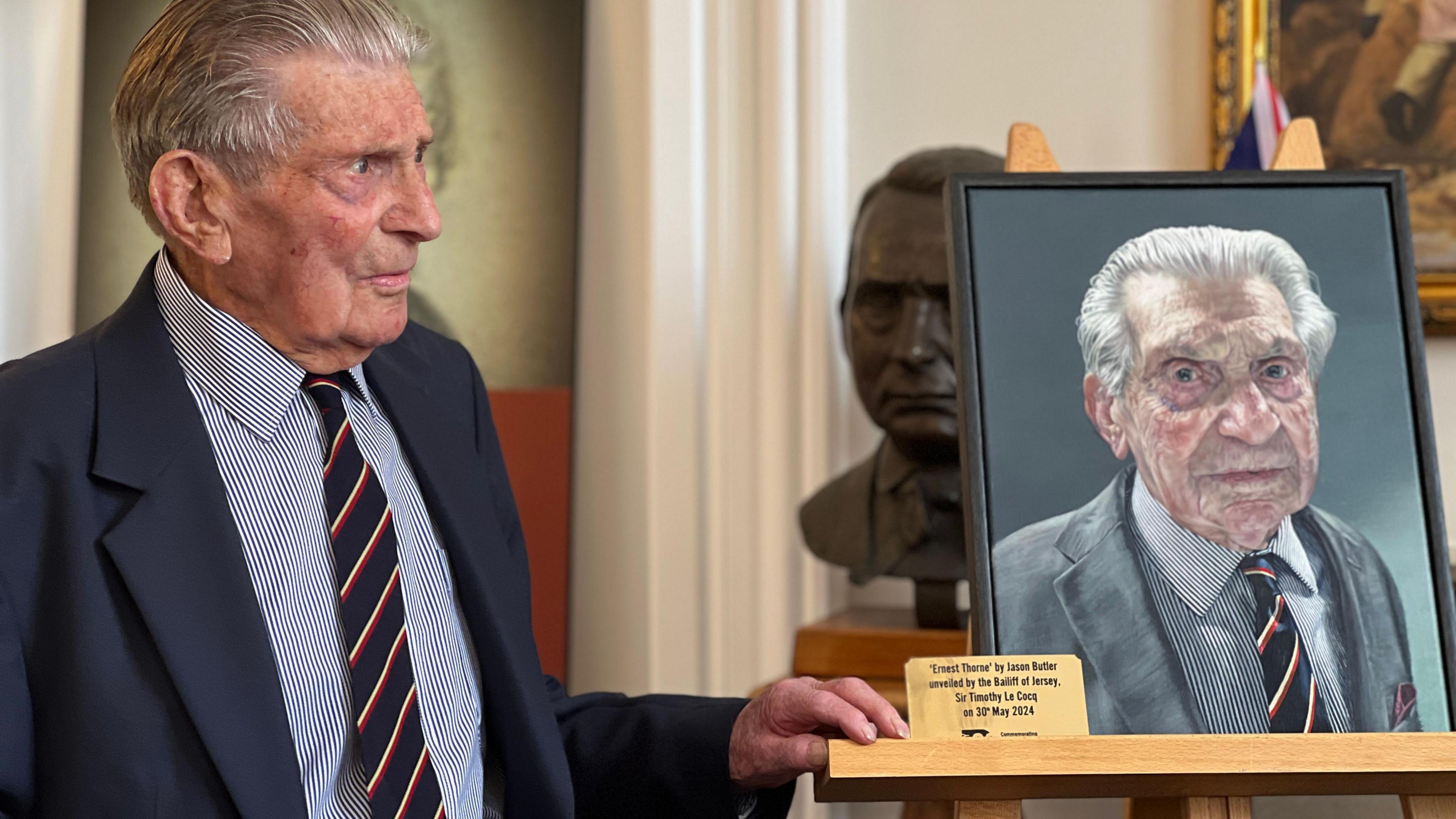 Ernest stands next to the portrait of himself and places one hand on the frame, he's wearing the same clothes as he is wearing in the portrait