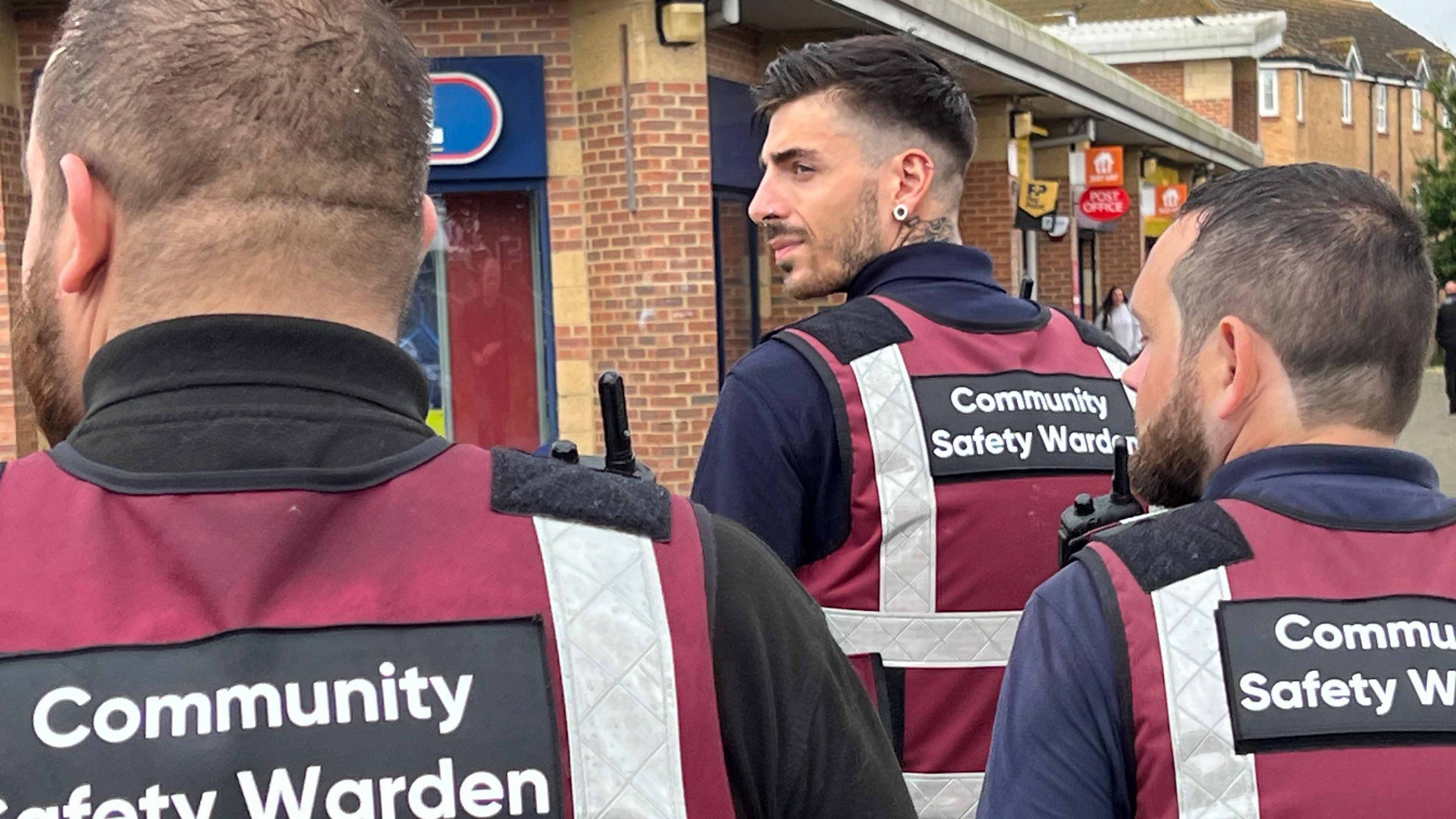 Three safety wardens photographed from behind wearing red vests that say Community Safety Warden on them