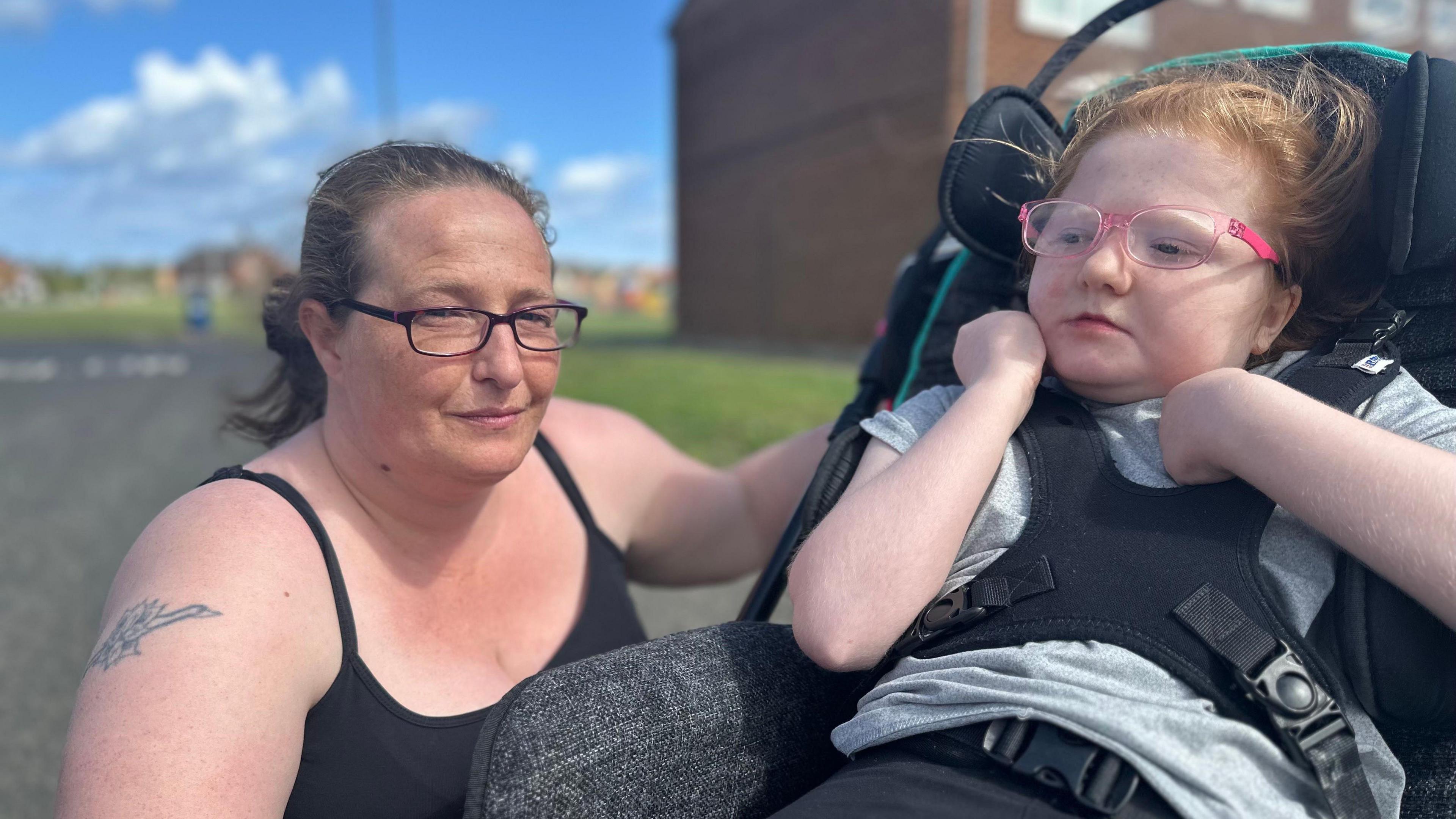 Gemma Wilkinson is kneeling next to her daughter Sarah's wheelchair. Gemma has brown hair, which is in a ponytail, and is wearing glasses. Sarah is strapped into the chair with a black harness. She has ginger hair and pink glasses.