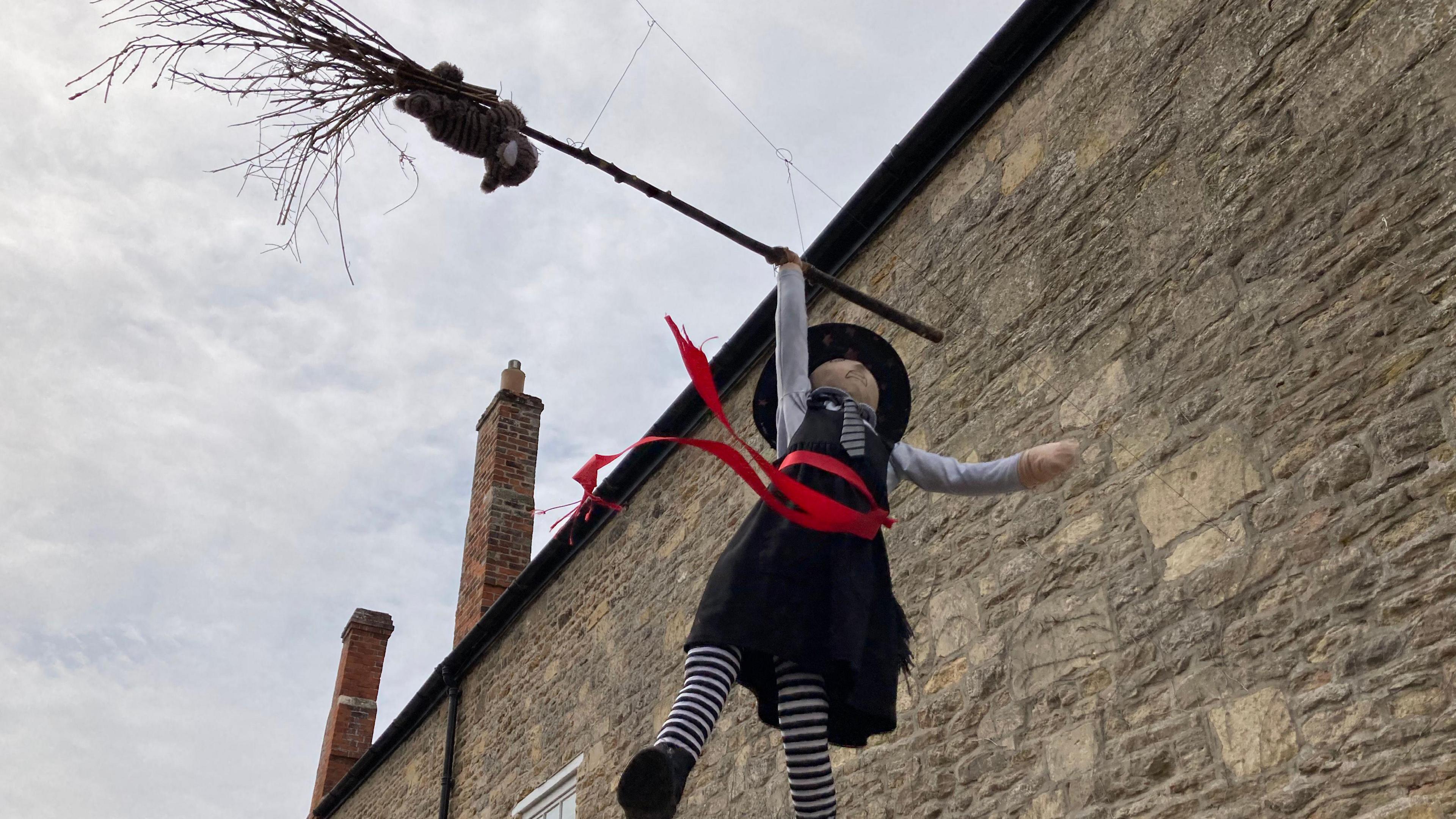 A scarecrow of a witch. She is wearing a black pointy hat, a black dress and striped tights. She is reaching up to grab a broomstick.