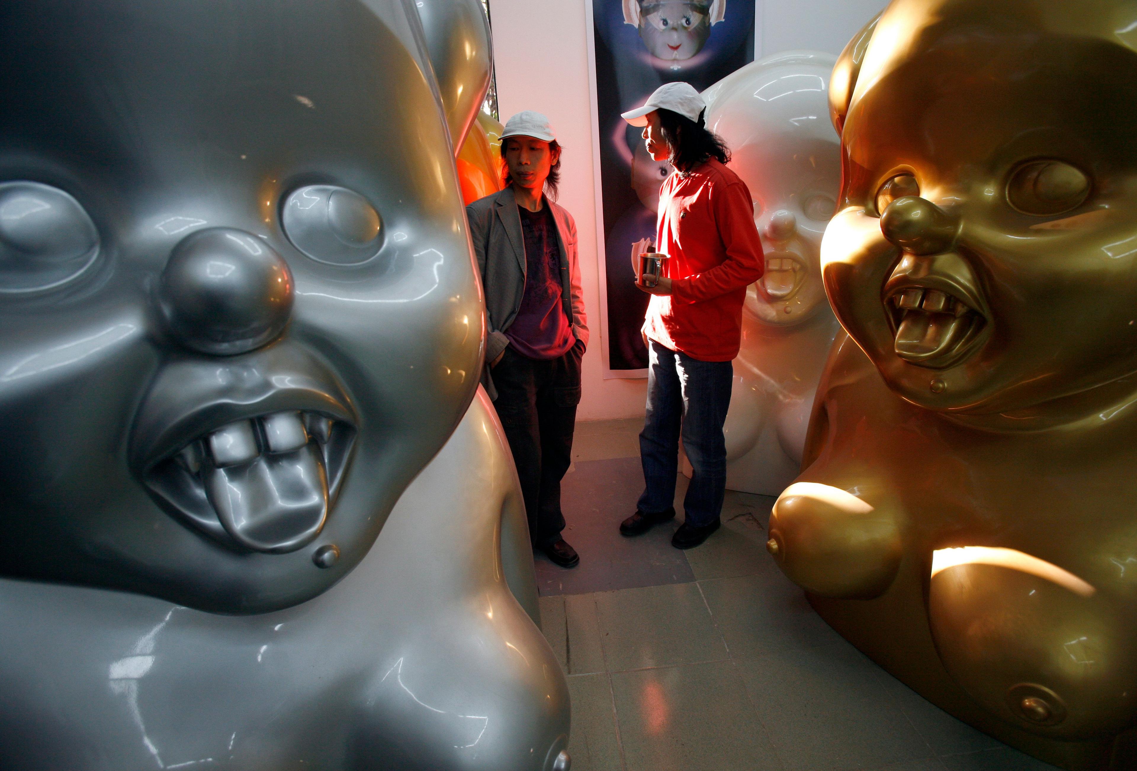 The Gao Brothers, both wearing white caps, stand between three of their "Miss Mao" sculptures. The sculptures are silver, gold and white and show a caricature-like Mao Zedong with buck-teeth, breasts and their tongues sticking out.