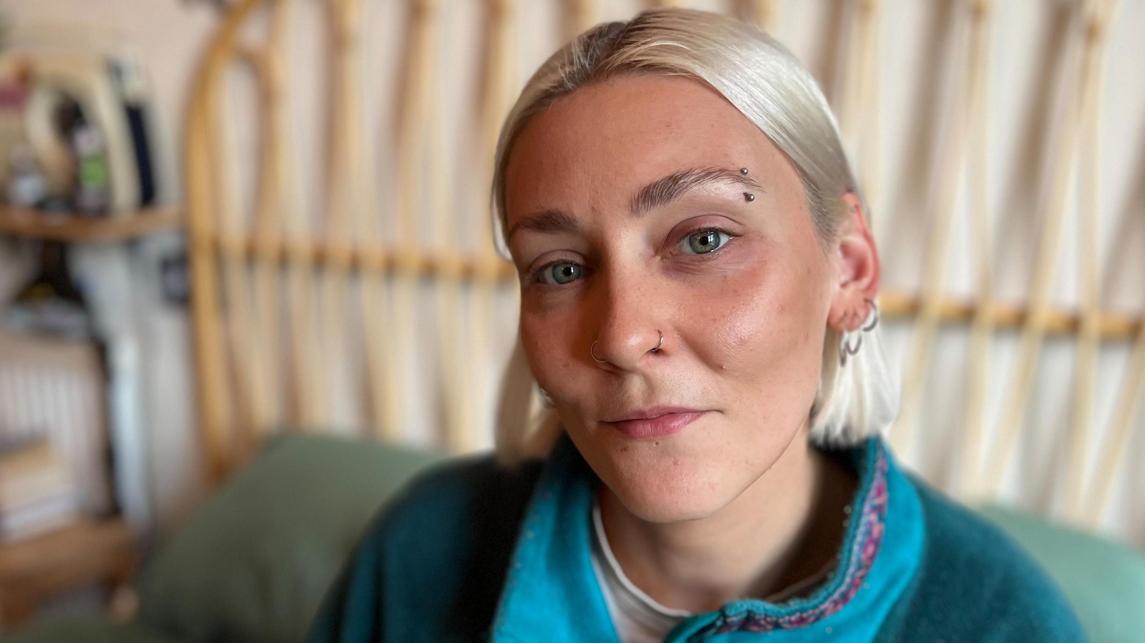 Elle Glenny, who has bobbed blond hair, looking directly at the camera and wearing a turquoise fleece. There is a bed frame in the background.
