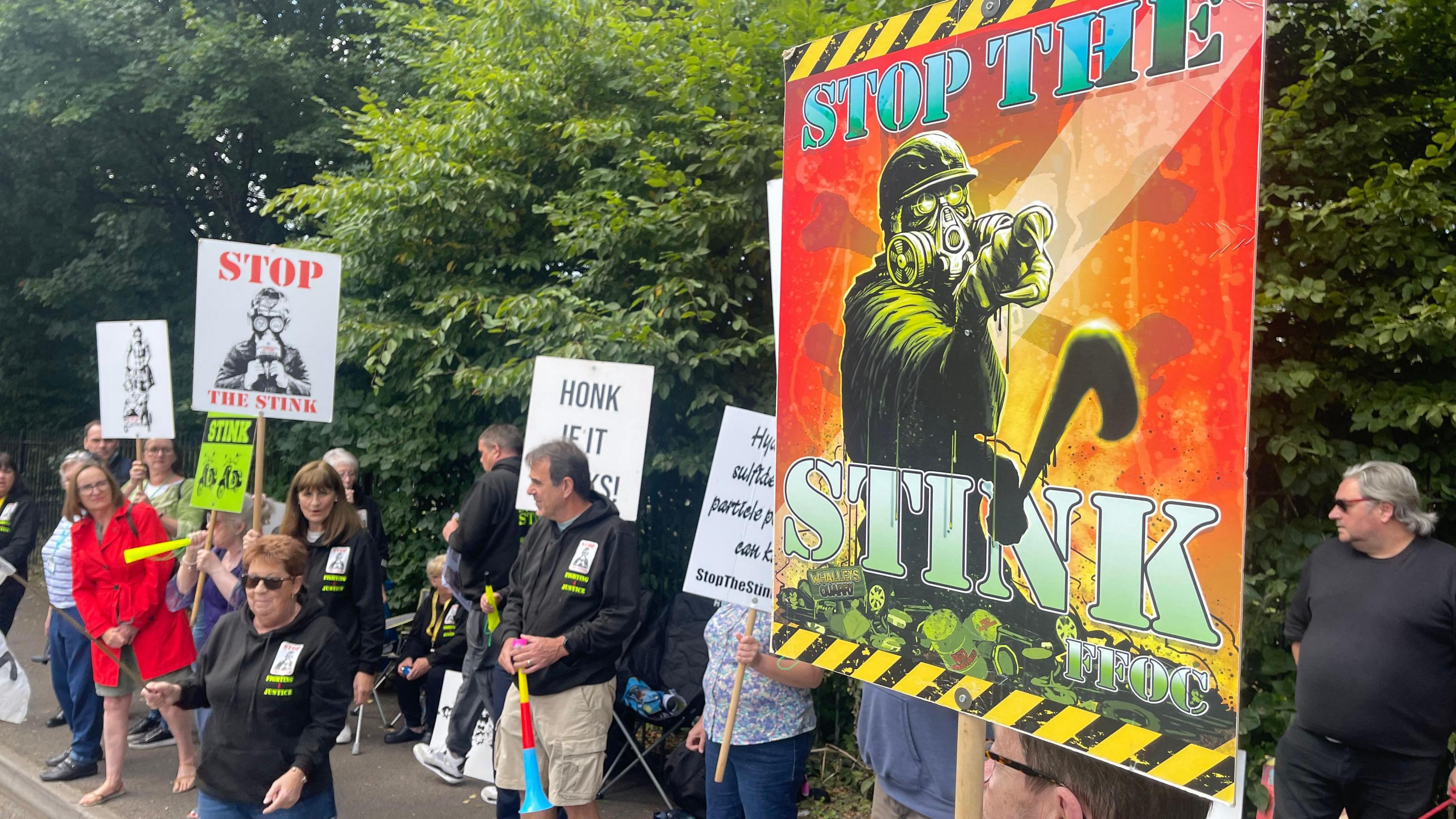 A group of people stand at the side of the road protesting about Walleys Quarry landfill. Several people are holding signs that say "stop the stink". 