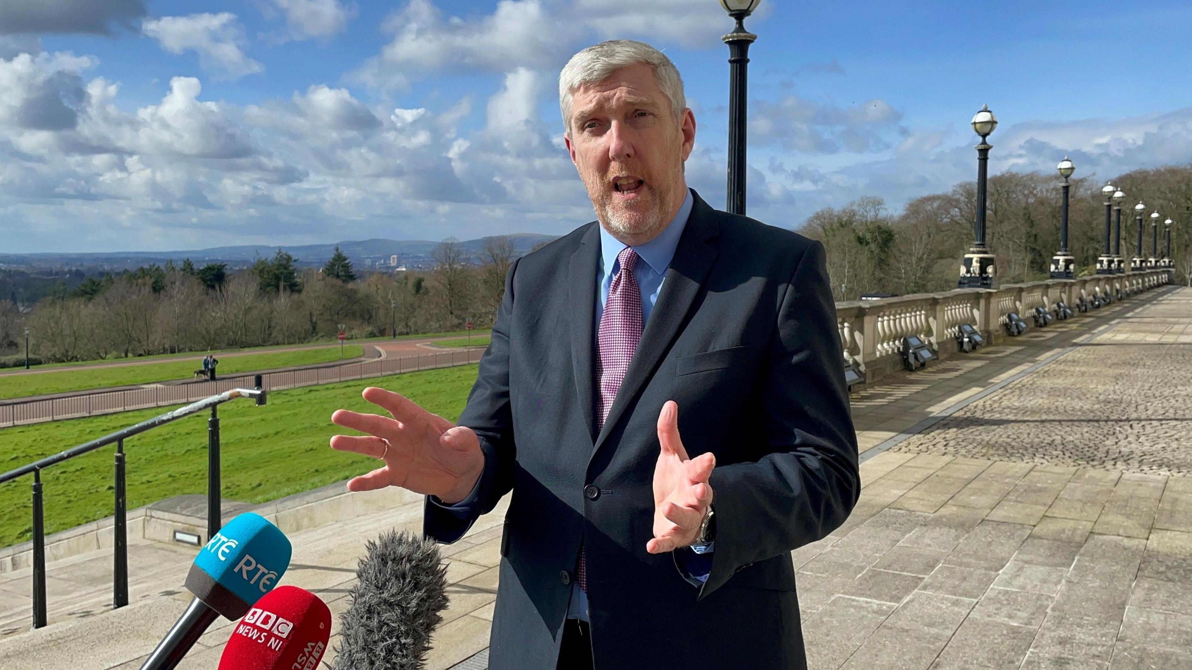 A man with white hair and a light-coloured beard is standing in front f three microphones and talking to someone off camera while gesturing with his hands. He is wearing a dark-coloured suit, a purple tie and a light blue suit. In the background you can see the Stormont estate including grey stones, steps and greenery. 