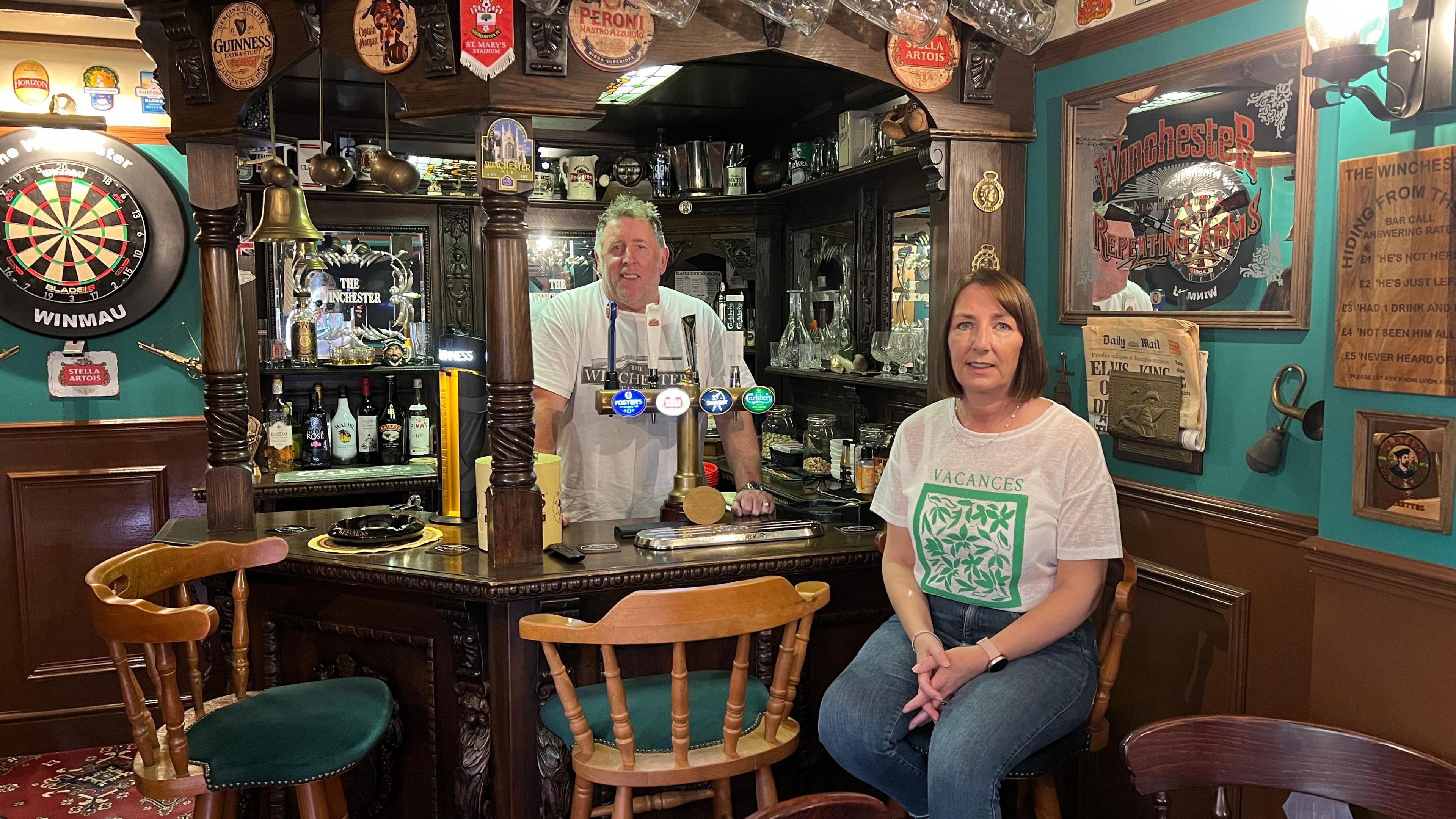 Kev Marchant standing behind his small bar of a little pub room with Gina Marchant sitting on a barstool in front of it. The walls are painted green and adorned with an assortment of items including beer mats, a dart board and horse brasses.