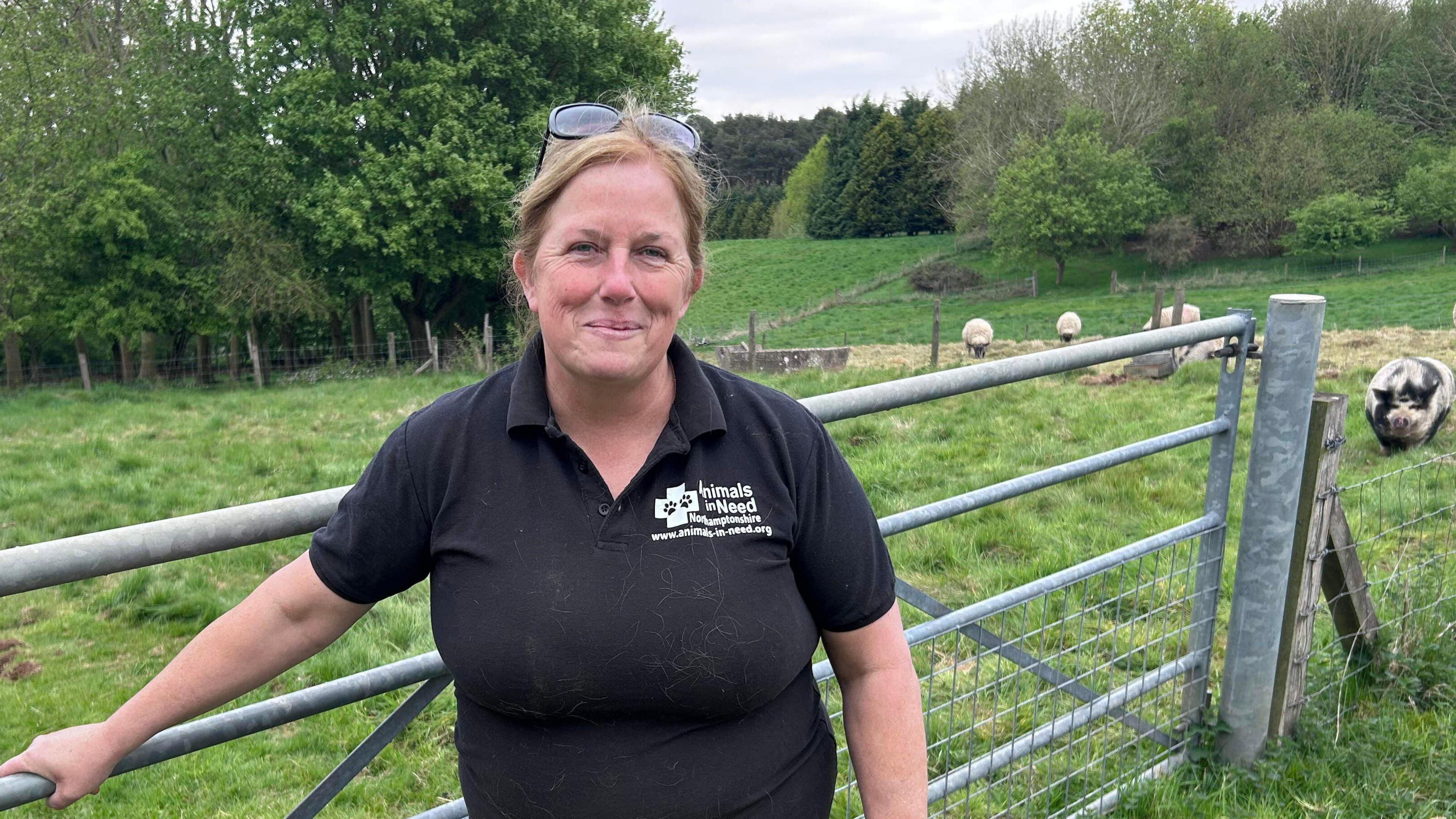 Woman in black t-shirt smiles in front of five bar gate. The background is a field of sheep and pigs.