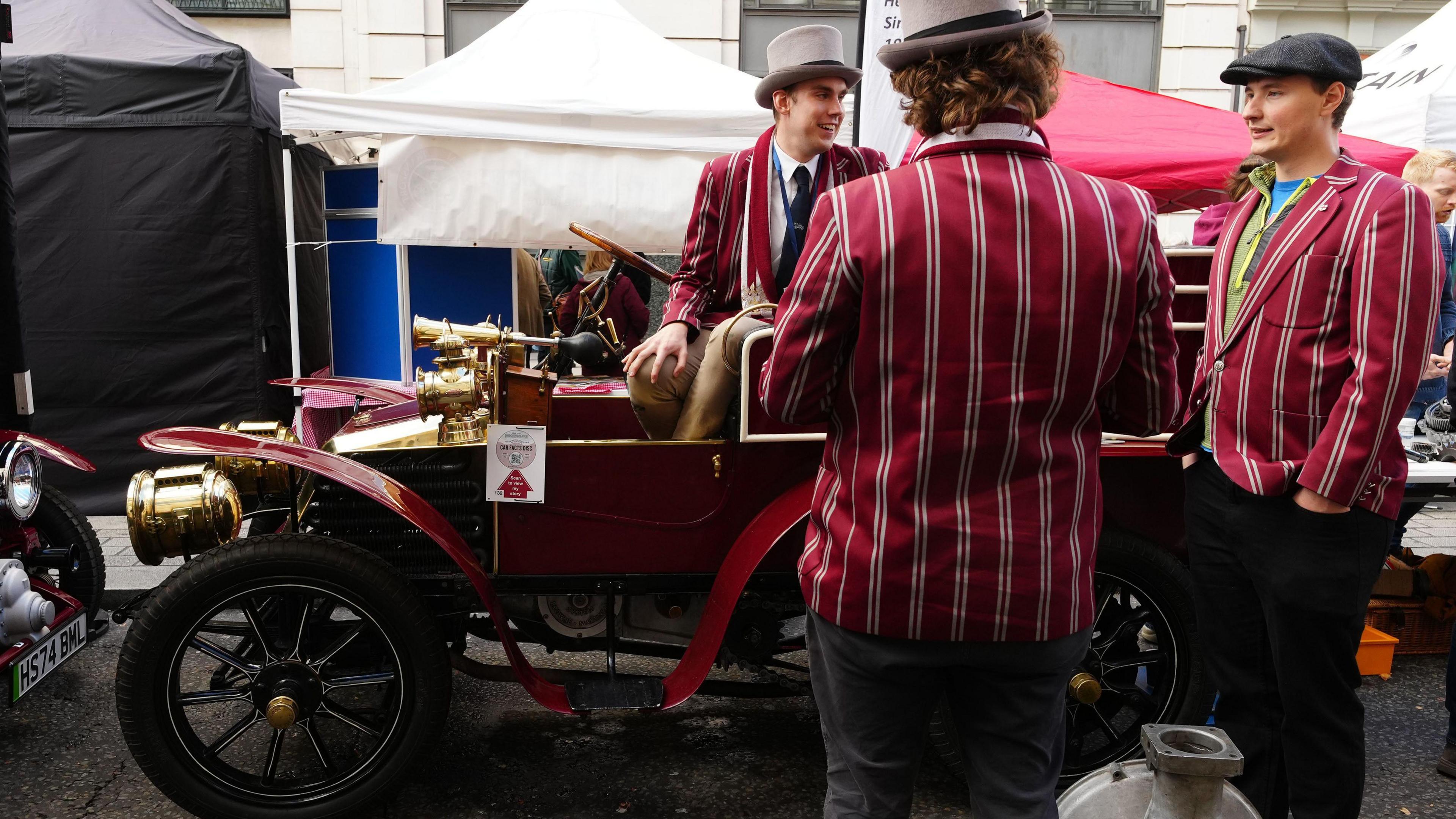 Three young men in stripy jackets surround an old car