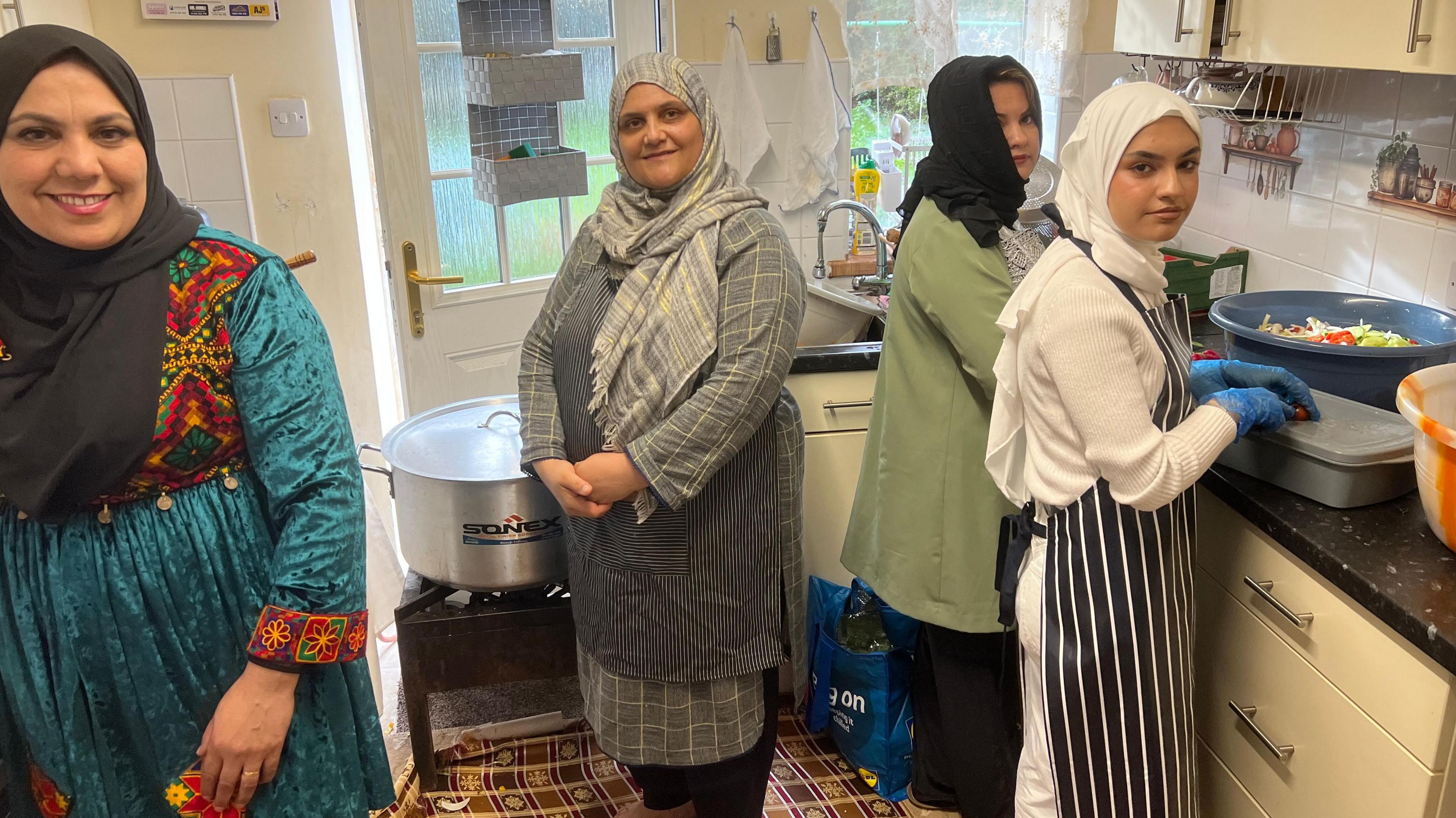 Four women stood in a kitchen area. The woman on the left is wearing a turquoise dress with embroidered patterns in red, yellow, and green, along with a black head covering. The second person is dressed in a grey striped tunic with a matching scarf. The third person is wearing a green jacket and a black head covering. The fourth person, on the far right, is wearing a white top, a black and white striped apron and blue gloves and is appearing to handle food in a large pan.