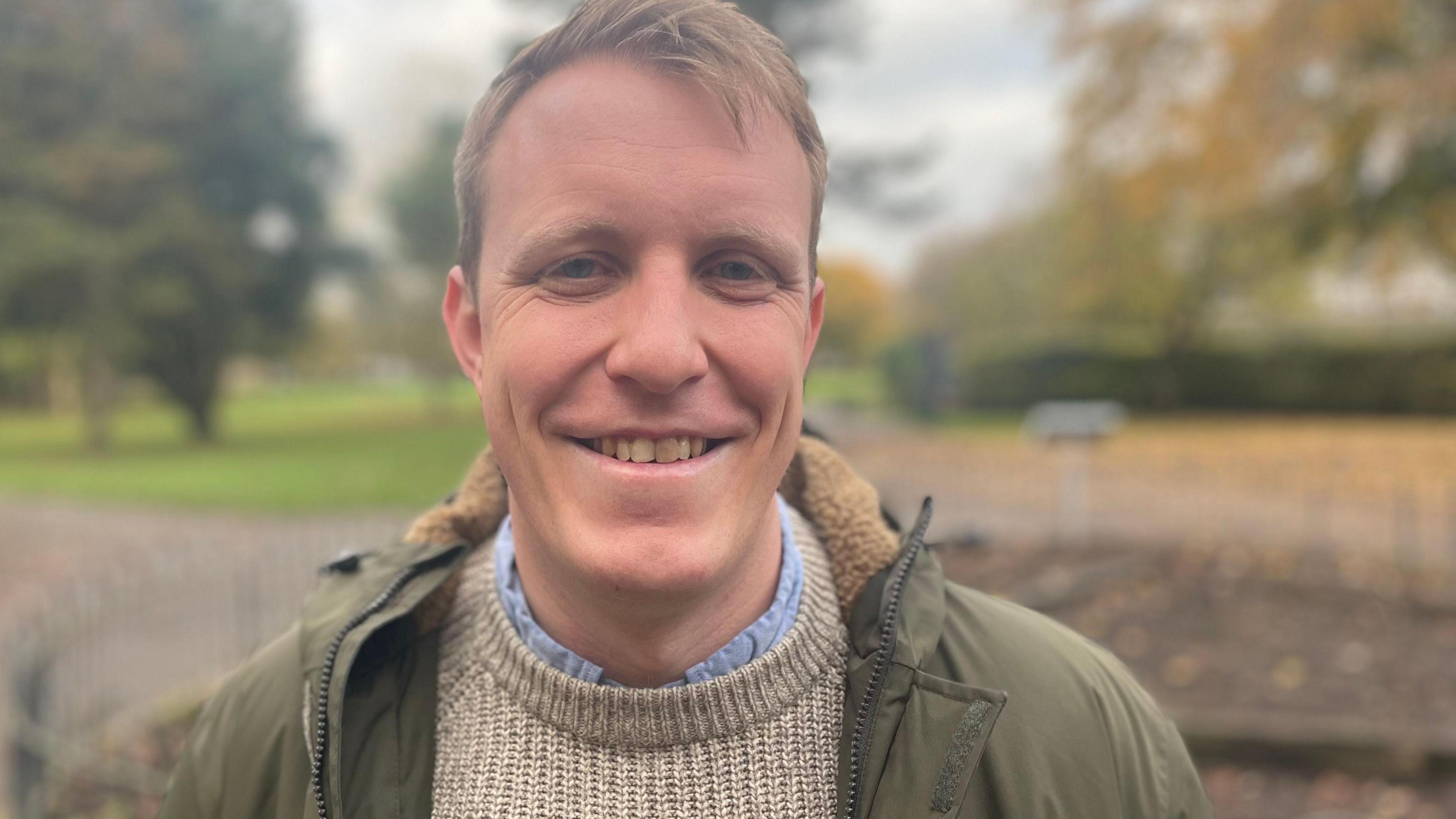 Dr Steffan Evans wearing a khaki coat, light jumper and blue shirt, smiling at the camera while standing in a park 