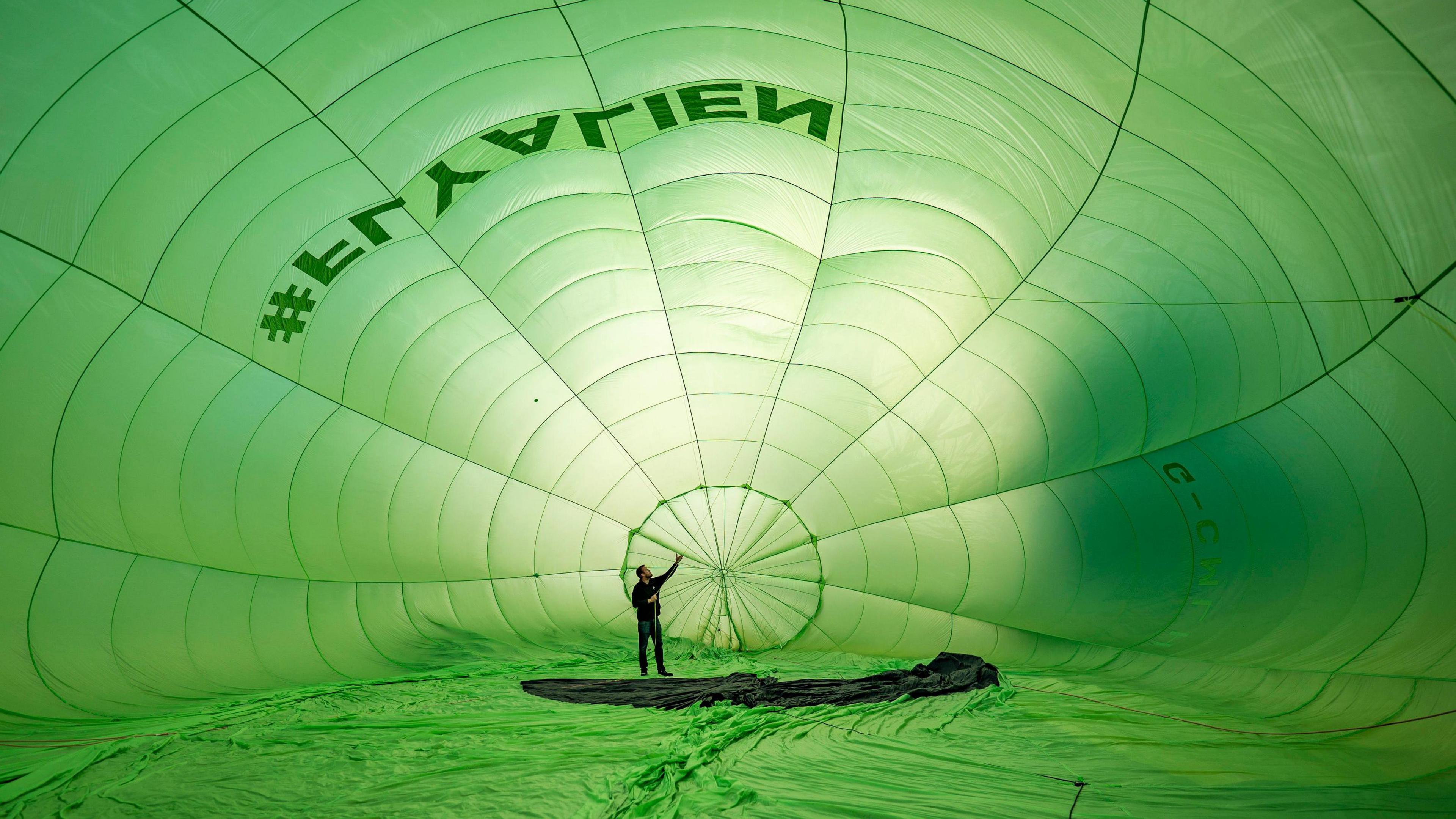 A man inside a light green hot air balloon. He is completely surrounded by the canopy and has one arm up, checking inside the balloon before it flies. 