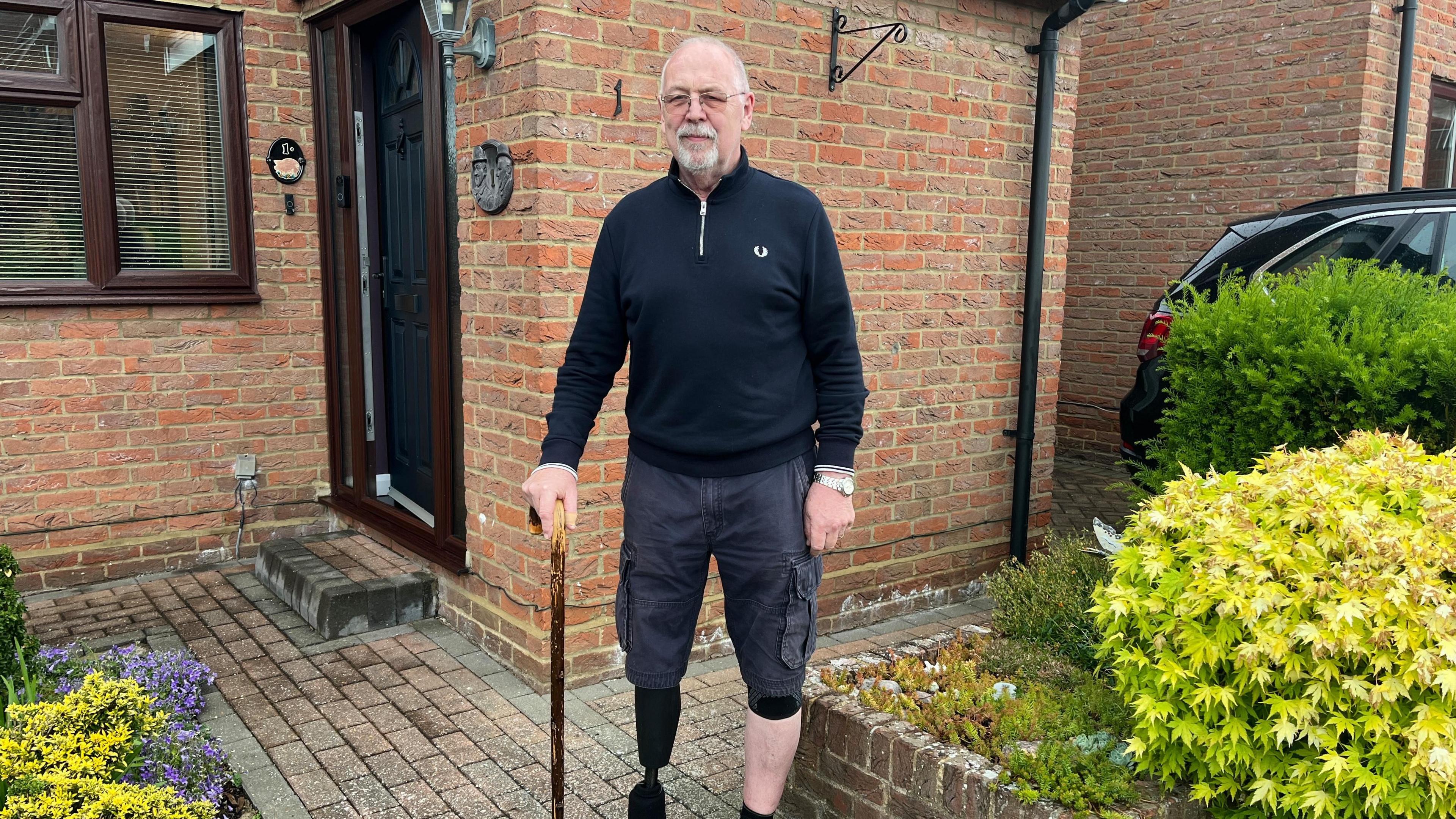 A man with a prosthetic leg standing outside a house with a walking stick. He is wearing a blue top with a zip and shorts. He has short grey hair and a short beard.