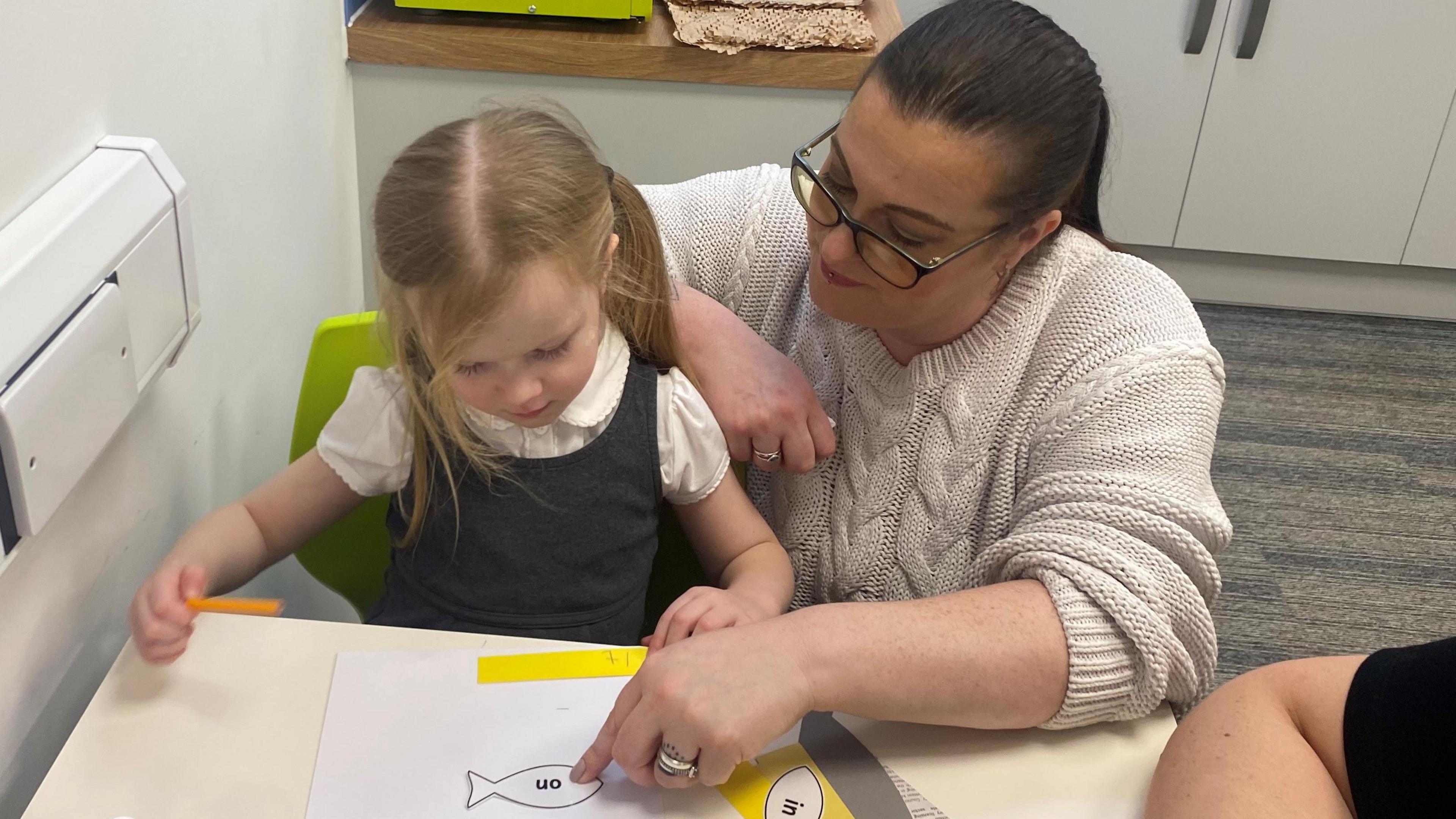 A young girl with pigtails wearing a grey school dress and white shirt. She is sat at a school table with her mum pointing to a sheet of paper on her desk, working on something together. Her mum has a black ponytail, glasses, and a beige jumper on.