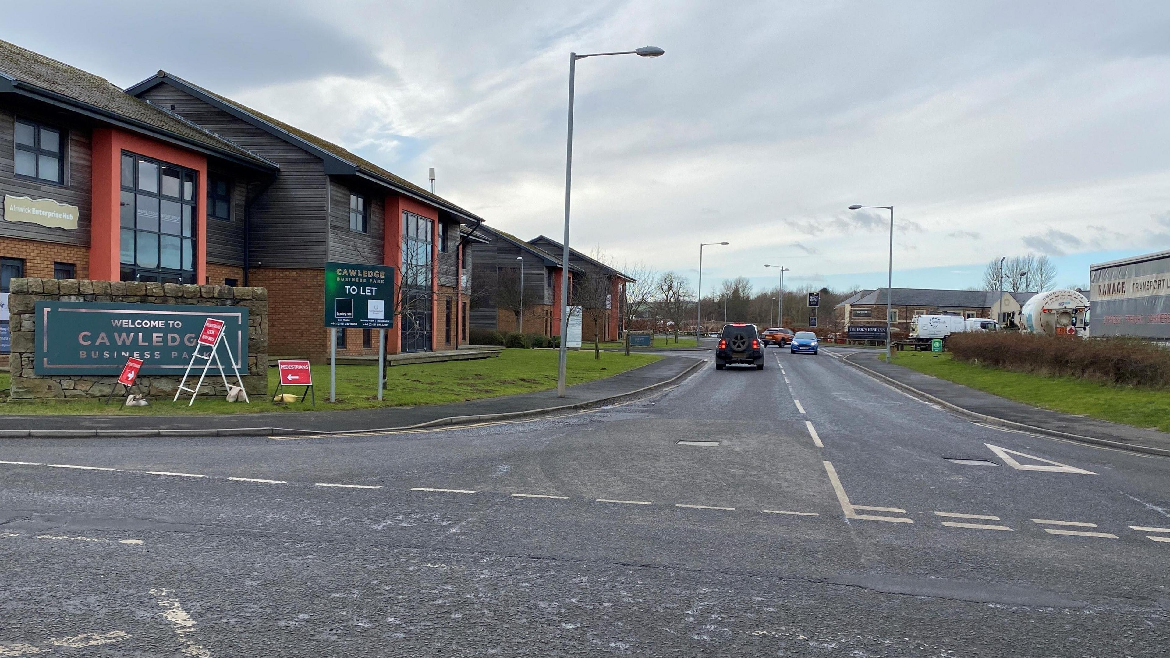 The entrance to the Cawledge Business Park with cars coming in and out. There is a sign for the park in the foreground and a pub called the Hogshead in the distance. There are buildings either side of the road.