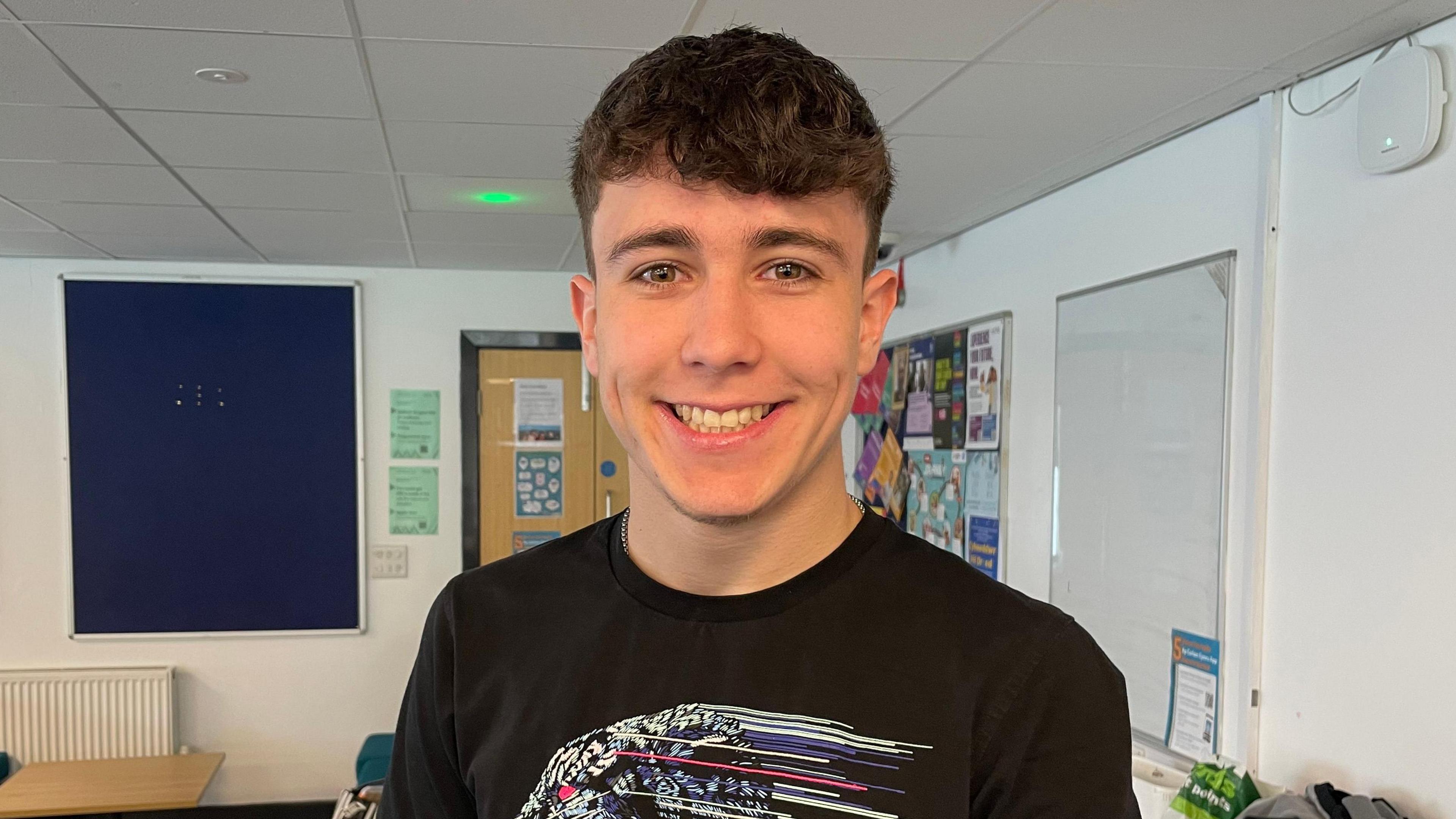 Dyn ifanc gyda gwallt byr brown yn gwisog crys-T du gyda phatrwm arno mewn lolfa chweched dosbarth mewn ysgol.