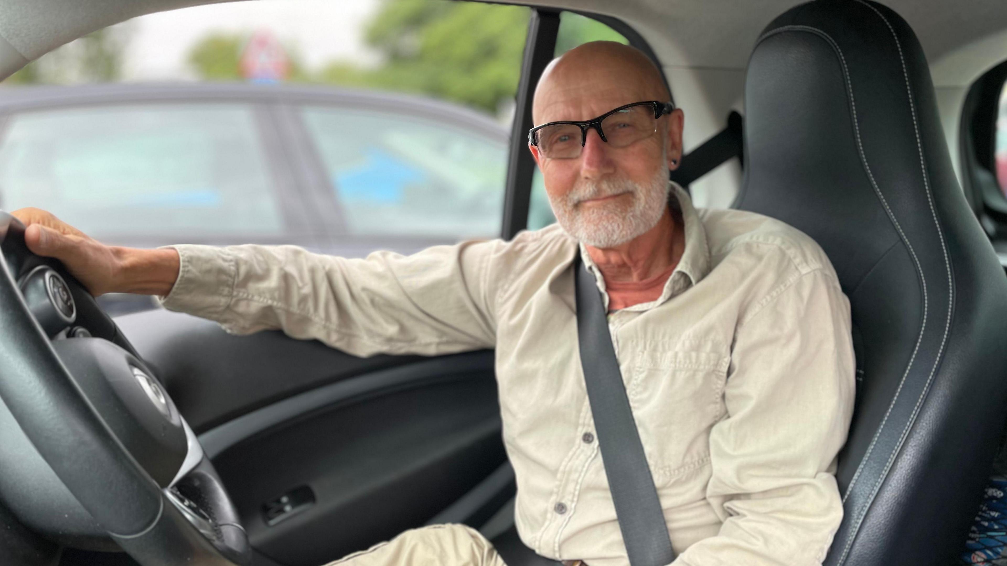A man sits in the driving seat of a car, with his right hand on the steering wheel and his seat belt on. He has a bald head and a white beard. He is wearing glasses and smiling at the camera. He is wearing a cream shirt.