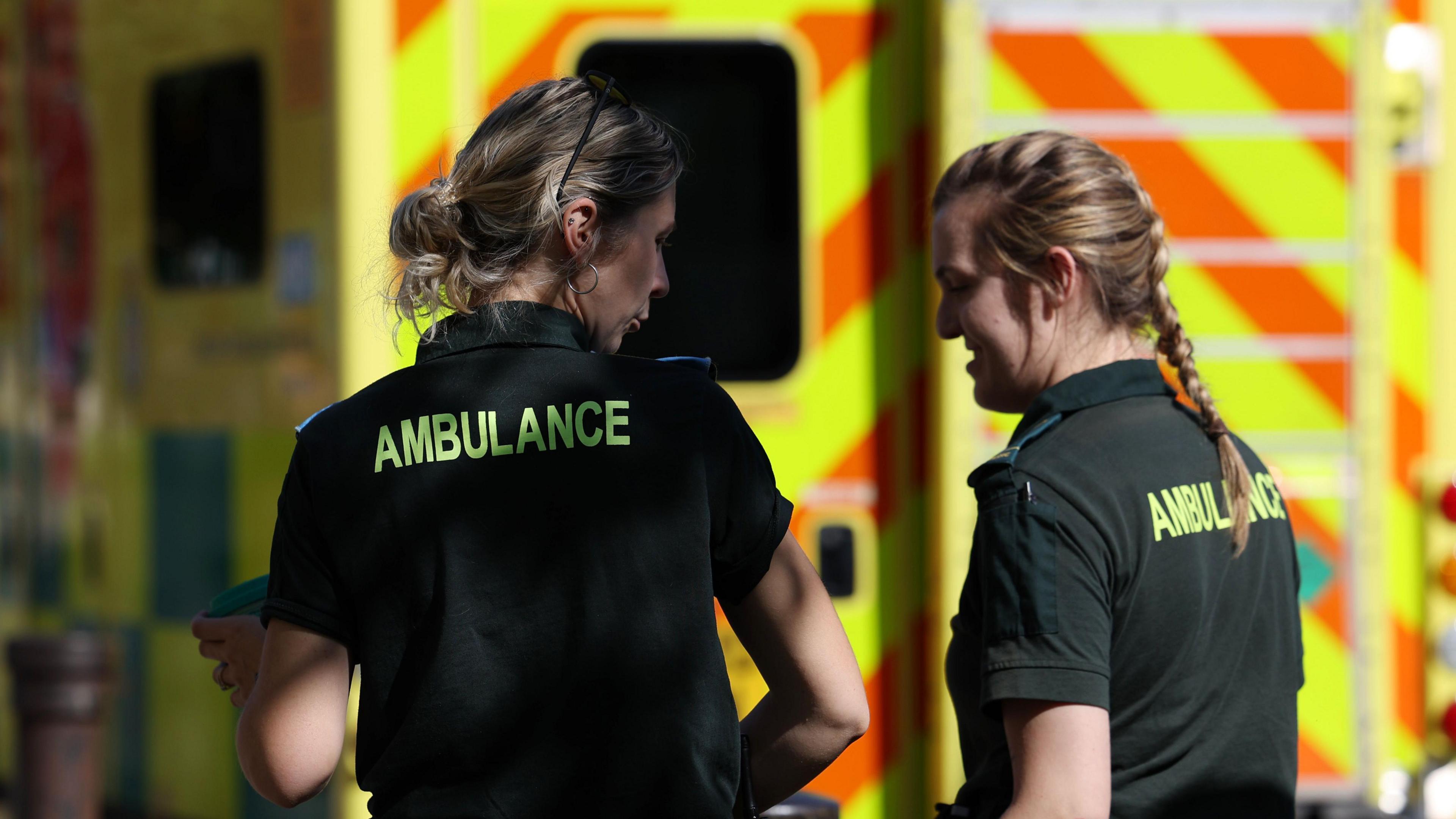 Two paramedics in green uniforms stand talking in front of an ambulance. 