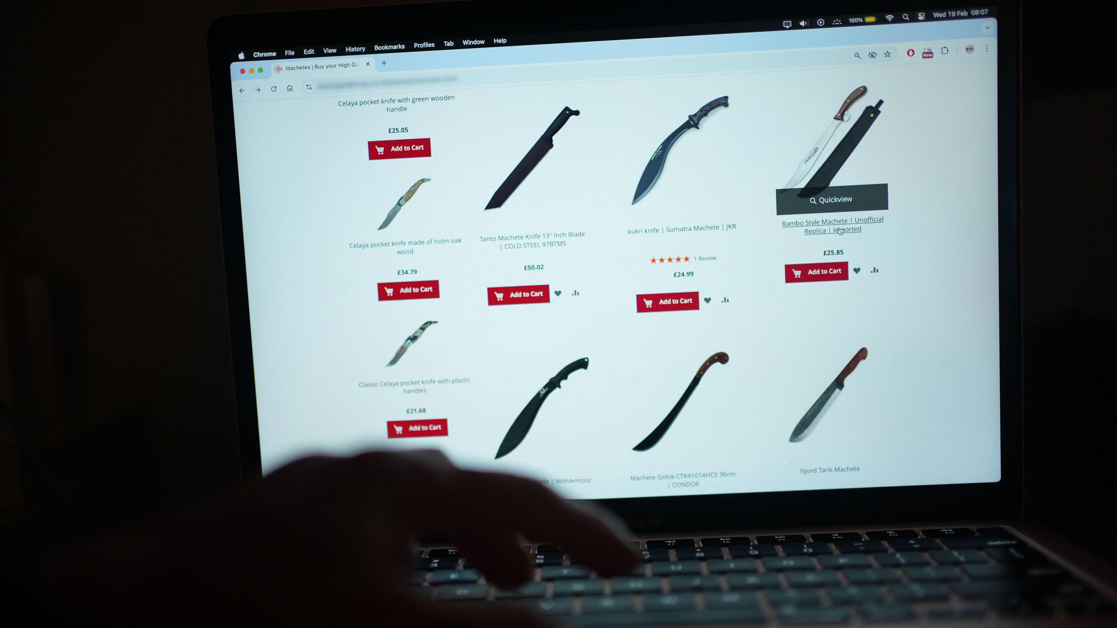 A website is open on a laptop displaying knives for sale. A person's hand is in the frame, hovering over the keyboard. The room behind the laptop is dark.