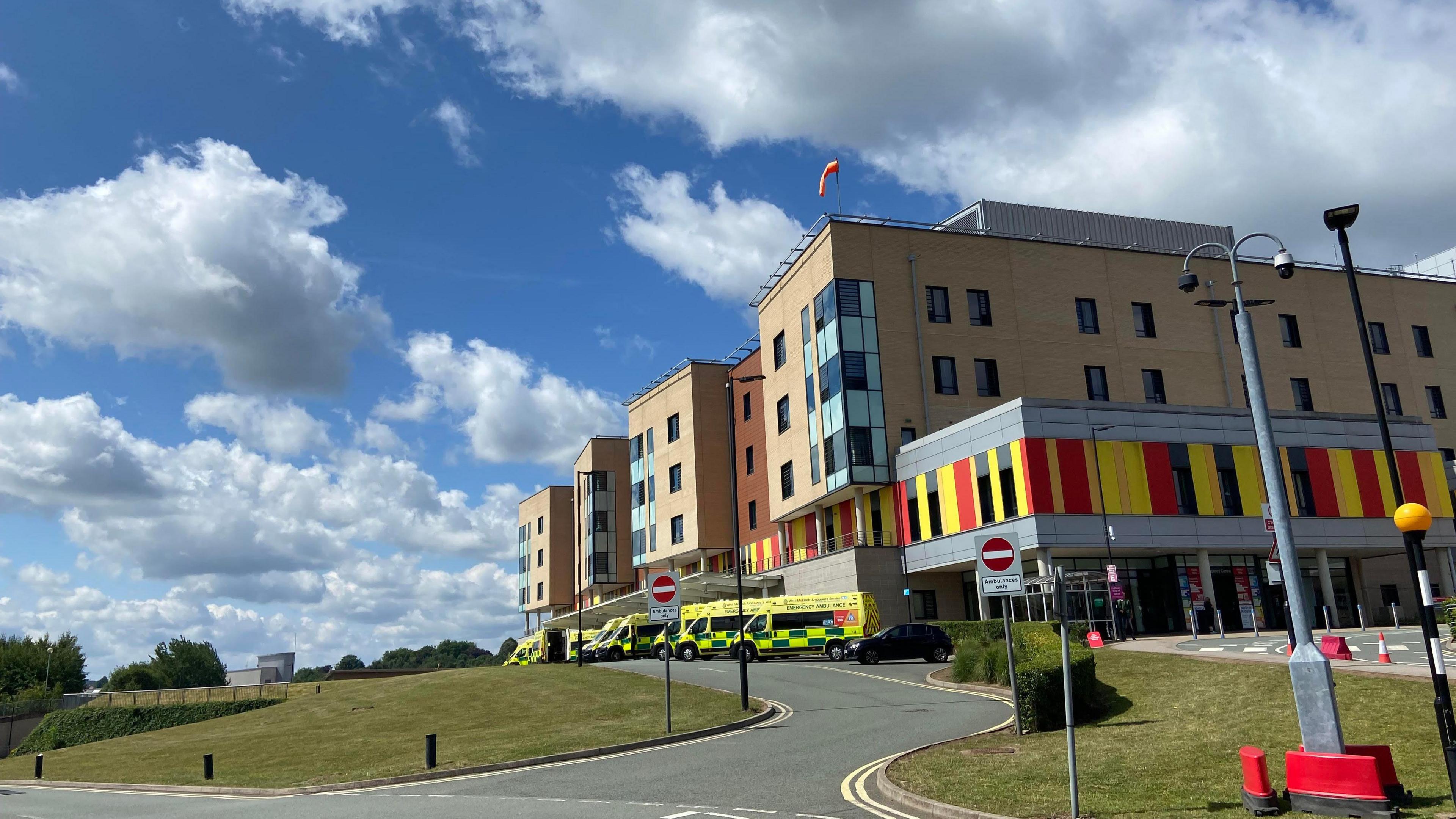 The outside of the Royal Stoke University Hospital, a five-storey building with red and yellow cladding. There are ambulances parked outside the building. There is a road leading up to the ambulance bays.