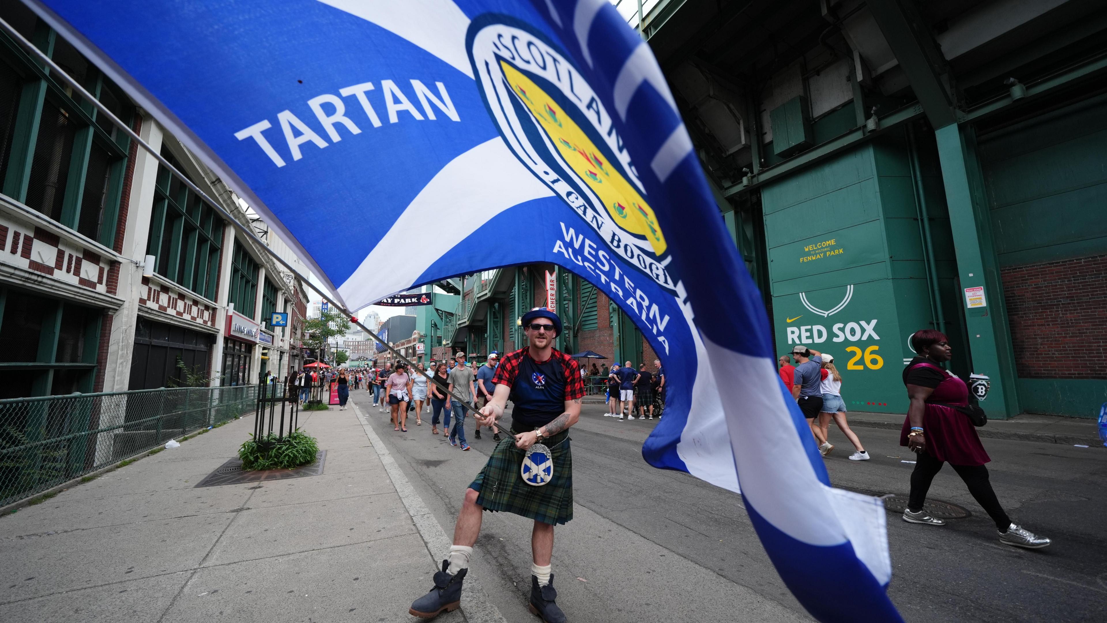 A Scotland fan in a kilt waves a large saltire flag outside the Boston Red Sox stadium