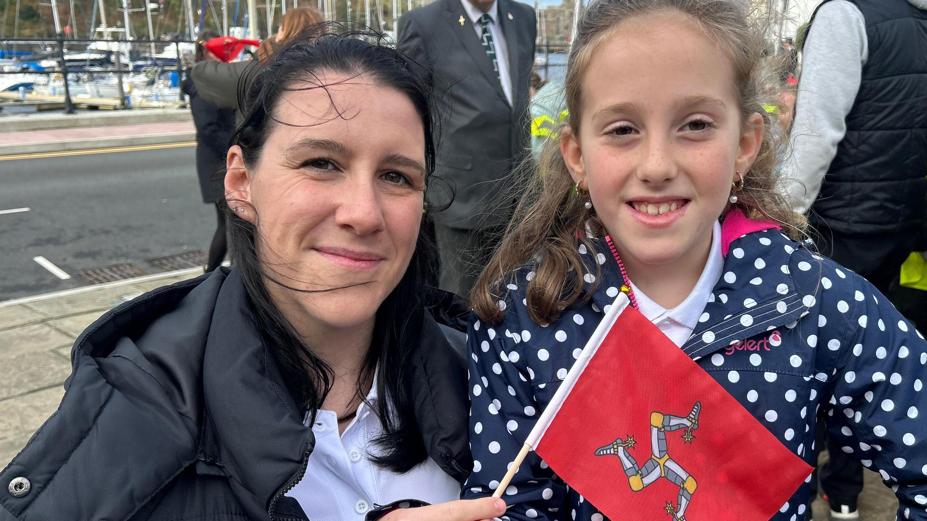 A woman and her nine year old daughter crouched down in front of a harbour marina. The mother has black, straight hair and a black outdoor coat, and is holding a small Isle of Man flag. The child is wearing a blue raincoat with white polka dots, and she has light brown hair. 