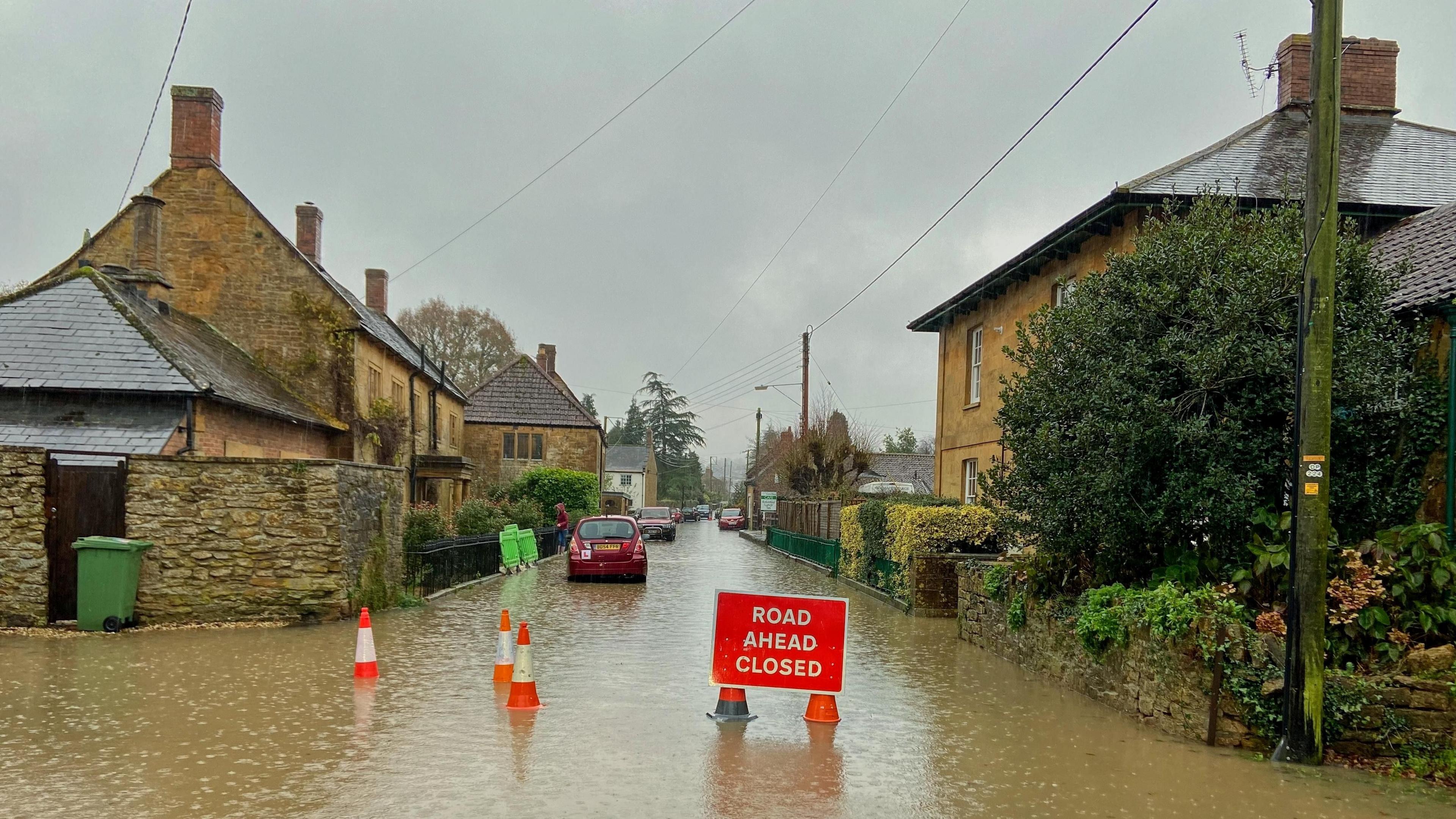 a flooded road in a village with parked cars, houses, a sign saying road ahead closed and three red and white transport cones.