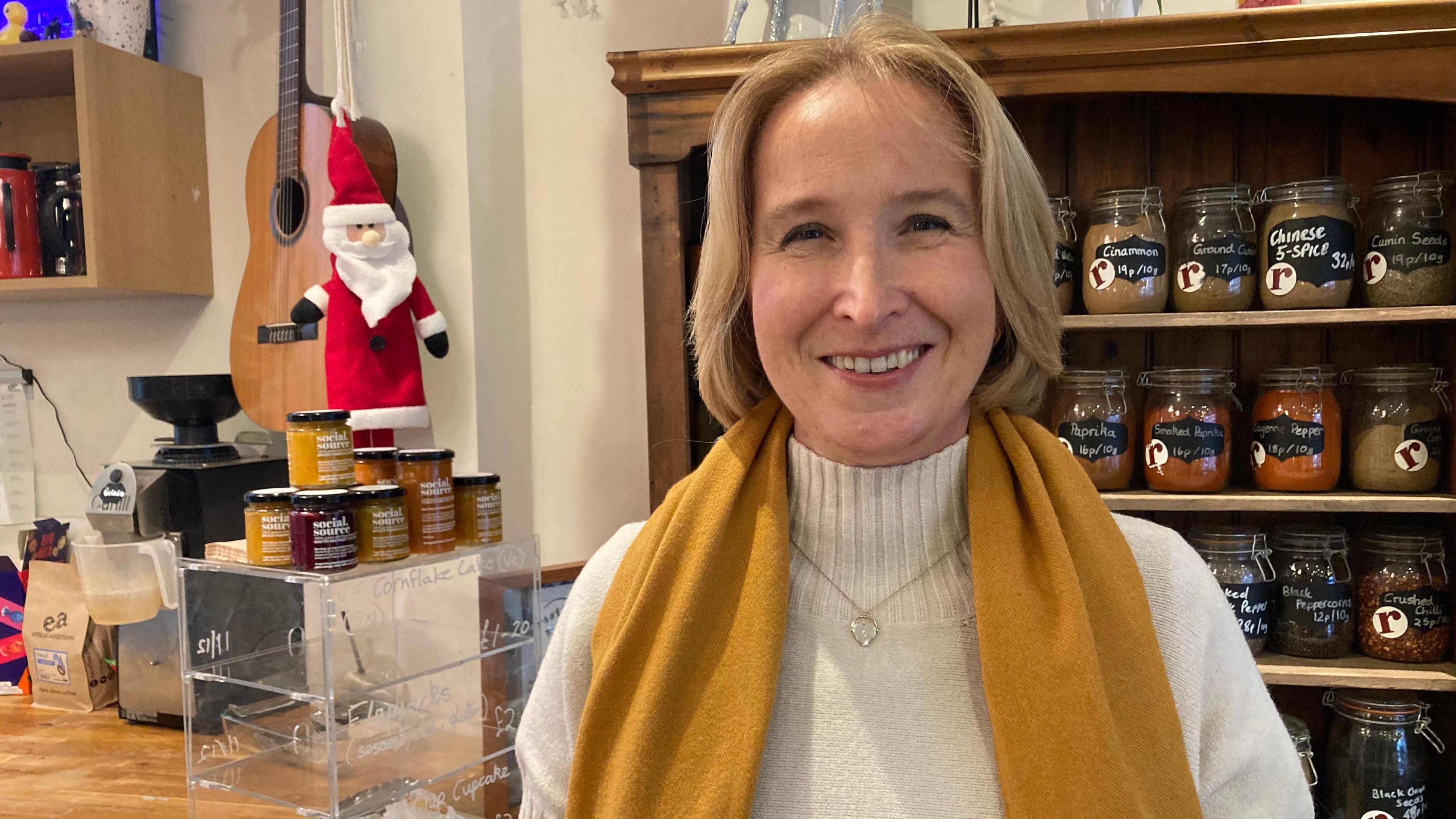 Claire Foote standing in the Roots cafe in front of a wooden dresser full of jars. She is wearing a cream high-neck jumper,  a heart shaped pendant necklace, and an orange scarf. She has blonde chin-length hair and is smiling at the camera. On her left is a Perspex display case with the jars of chutney stacked on top. 