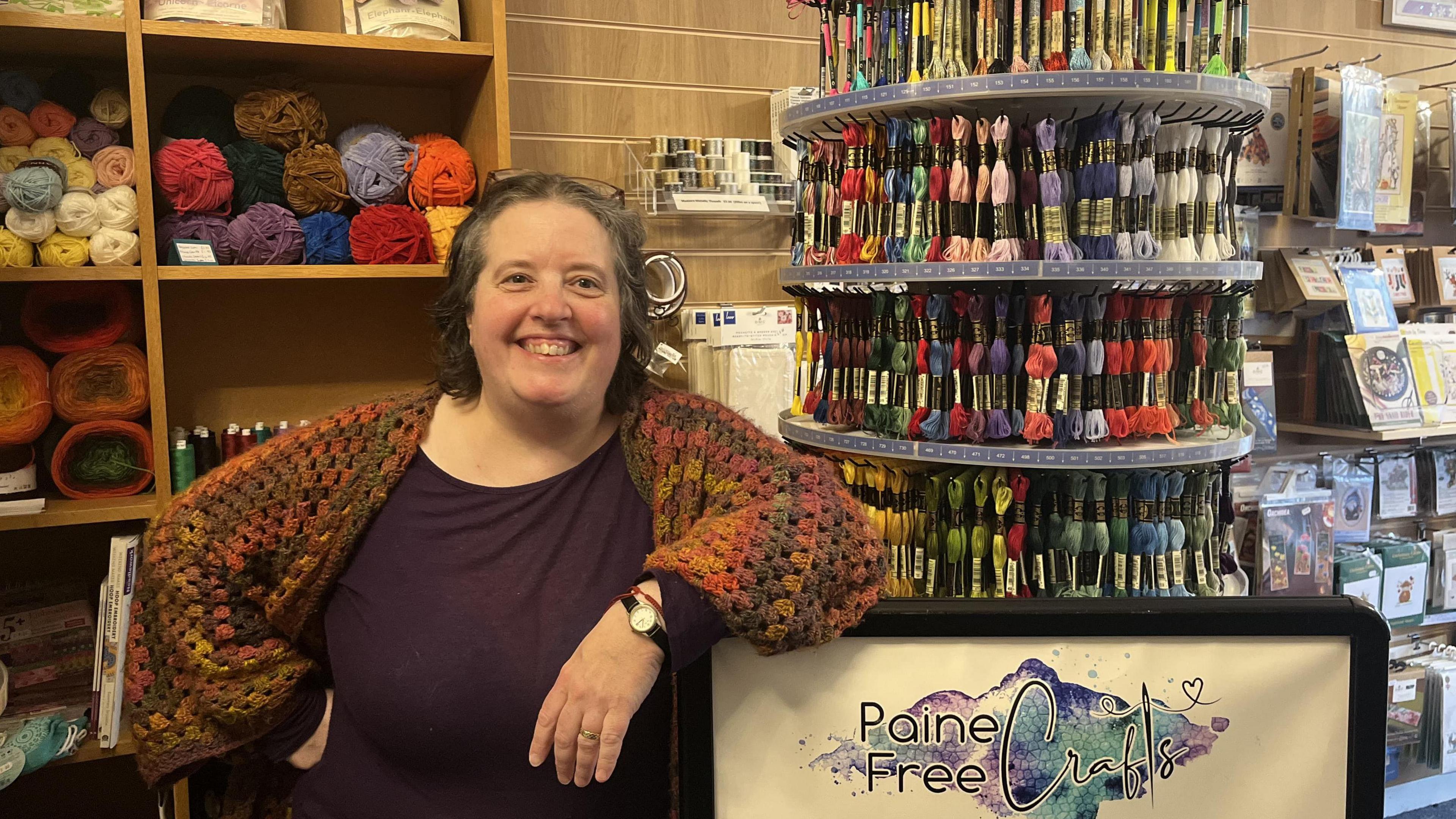 A woman with a rainbow coloured cardigan and purple t-shirt leans on a shop sign saying "Paine Free Crafts." The background shows balls of wool and craft equipment.