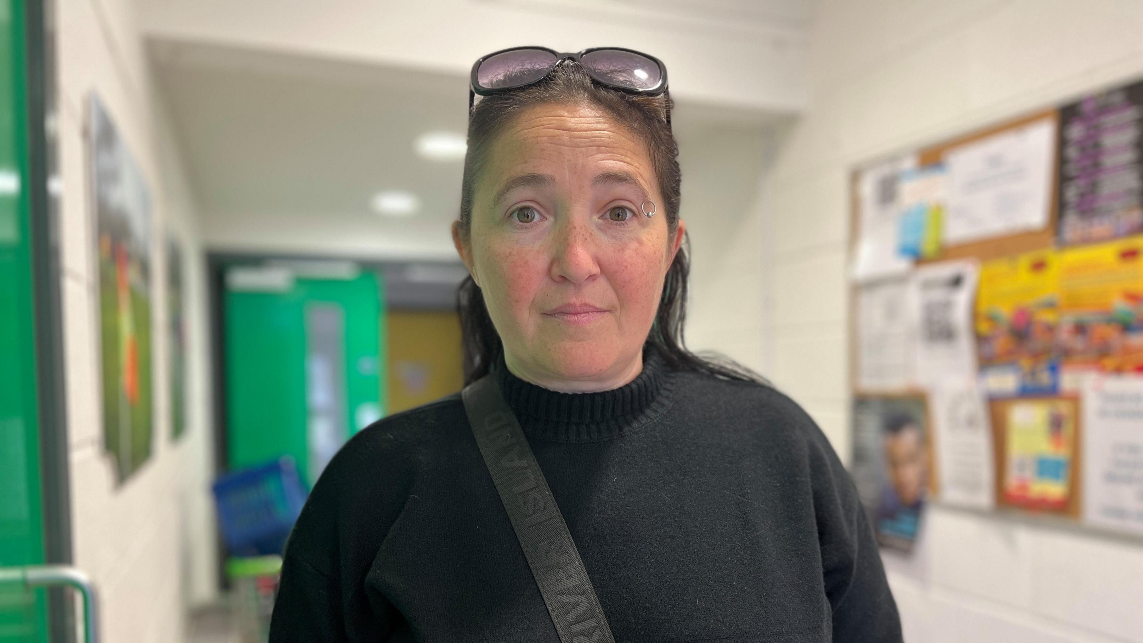Hayley Huntley wearing a black turtleneck jumper and large round sunglasses on her head. She has an eyebrow piercing and a black bag strap across her chest. she is standing in what looks like a school hallway, with noticeboards on the wall and bright green double doors.