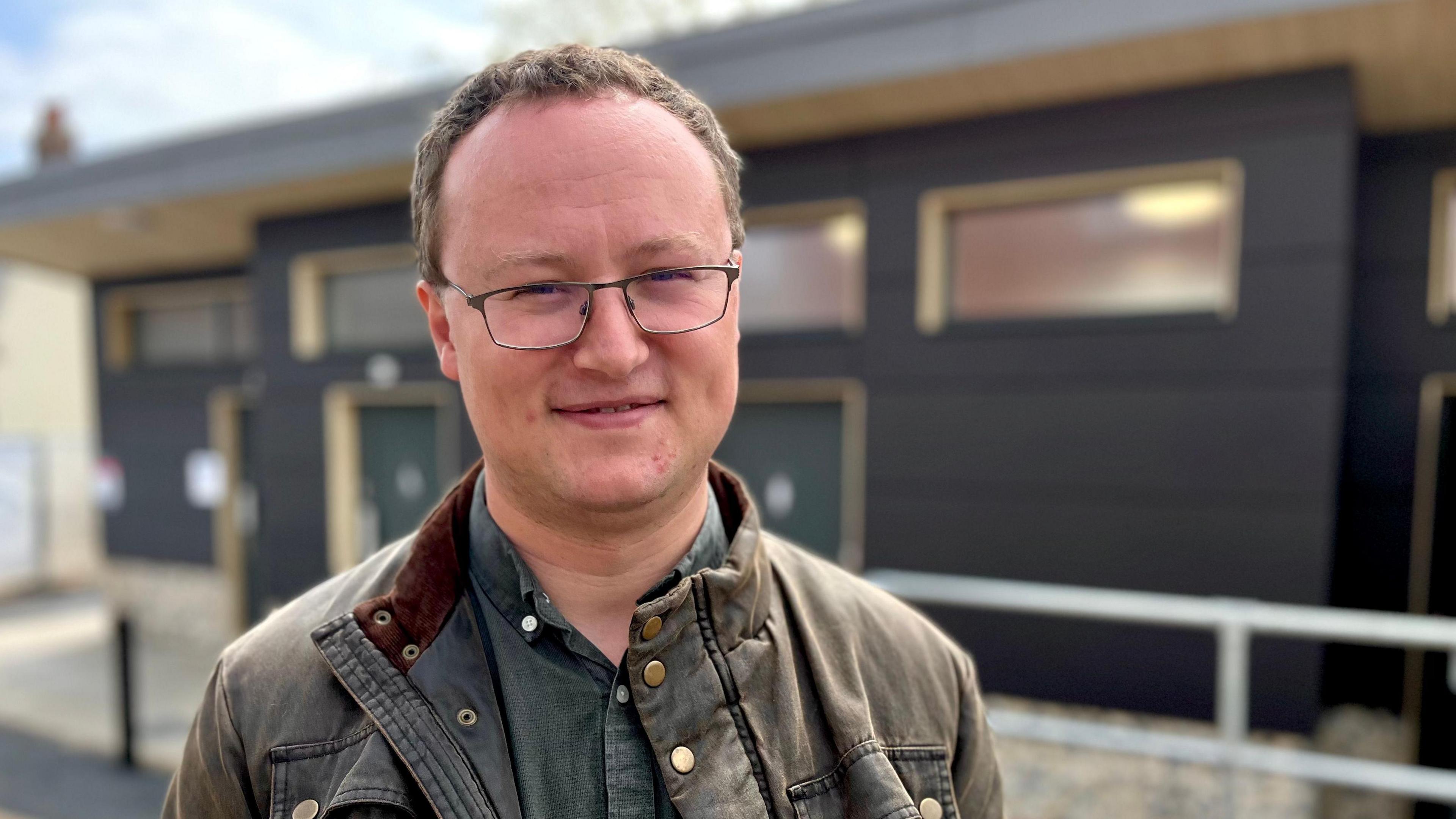 Tim Adams, standing in front of a building. He's wearing a green/brown jacket and glasses.