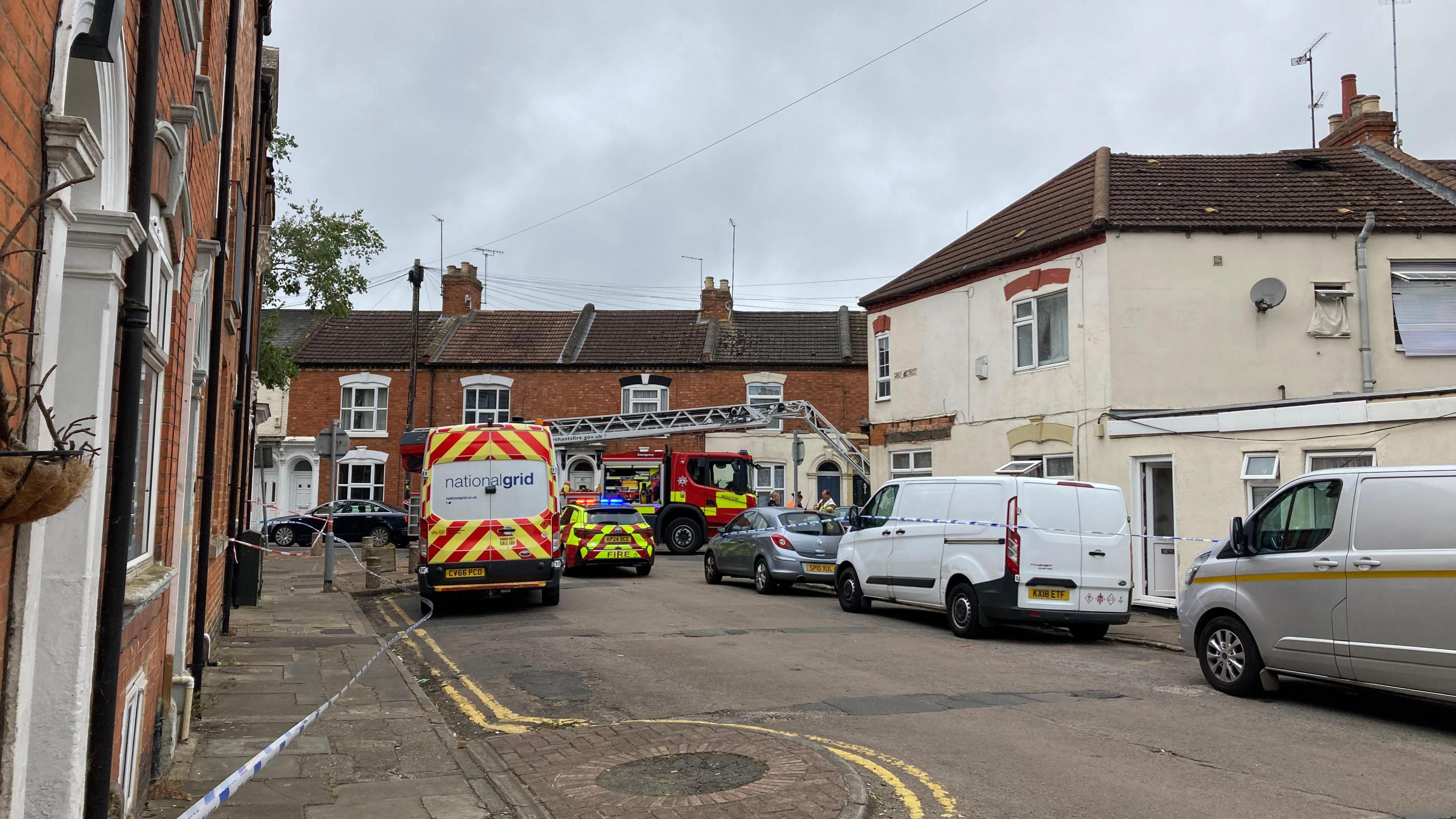 A fire engine, a police car and a national grid van parked in front of terraced houses with a blue police cordon in place.