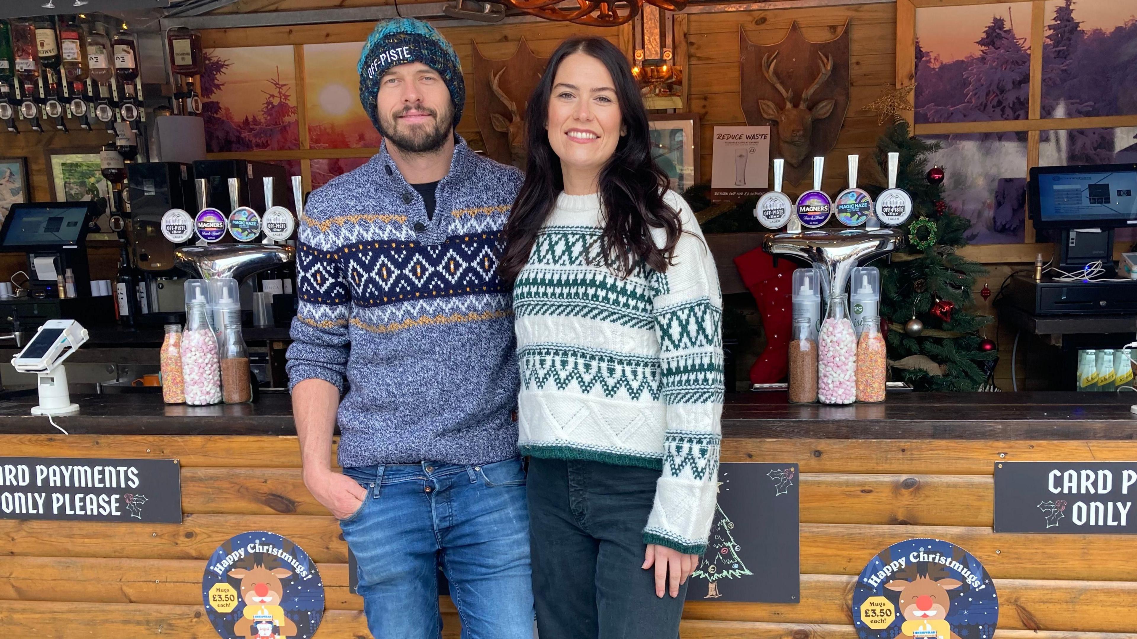 Abbey Matthews and her partner Kyle wearing Nordic style jumpers standing in front of one of their Bar At Yours stalls- they both have jeans on and are smiling in front of bar pumps. 