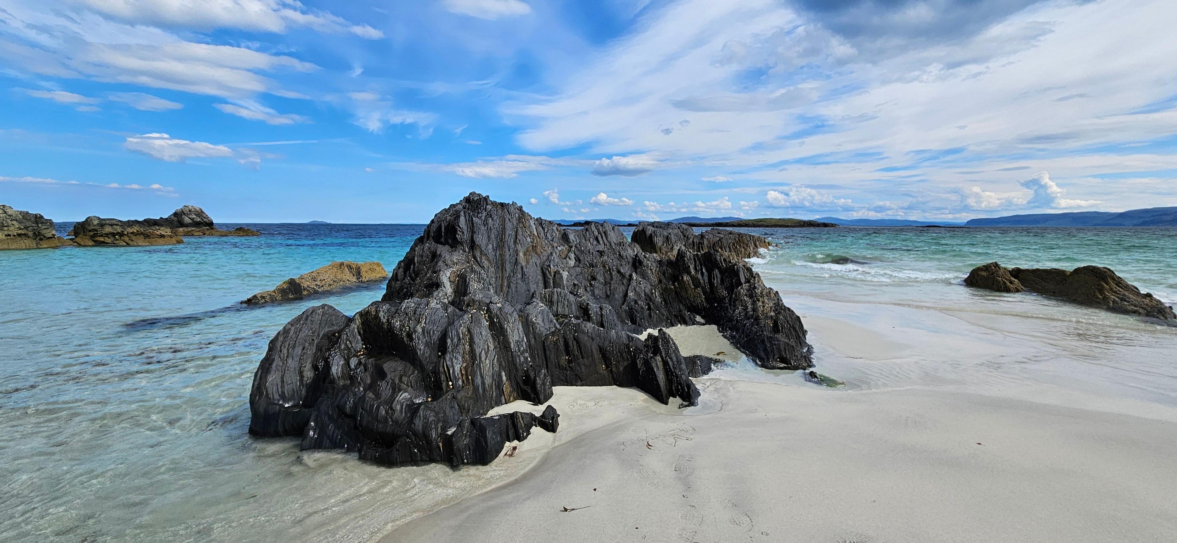 Landscape image of a large black rock on the beach with a blue ombre sea in the distance 