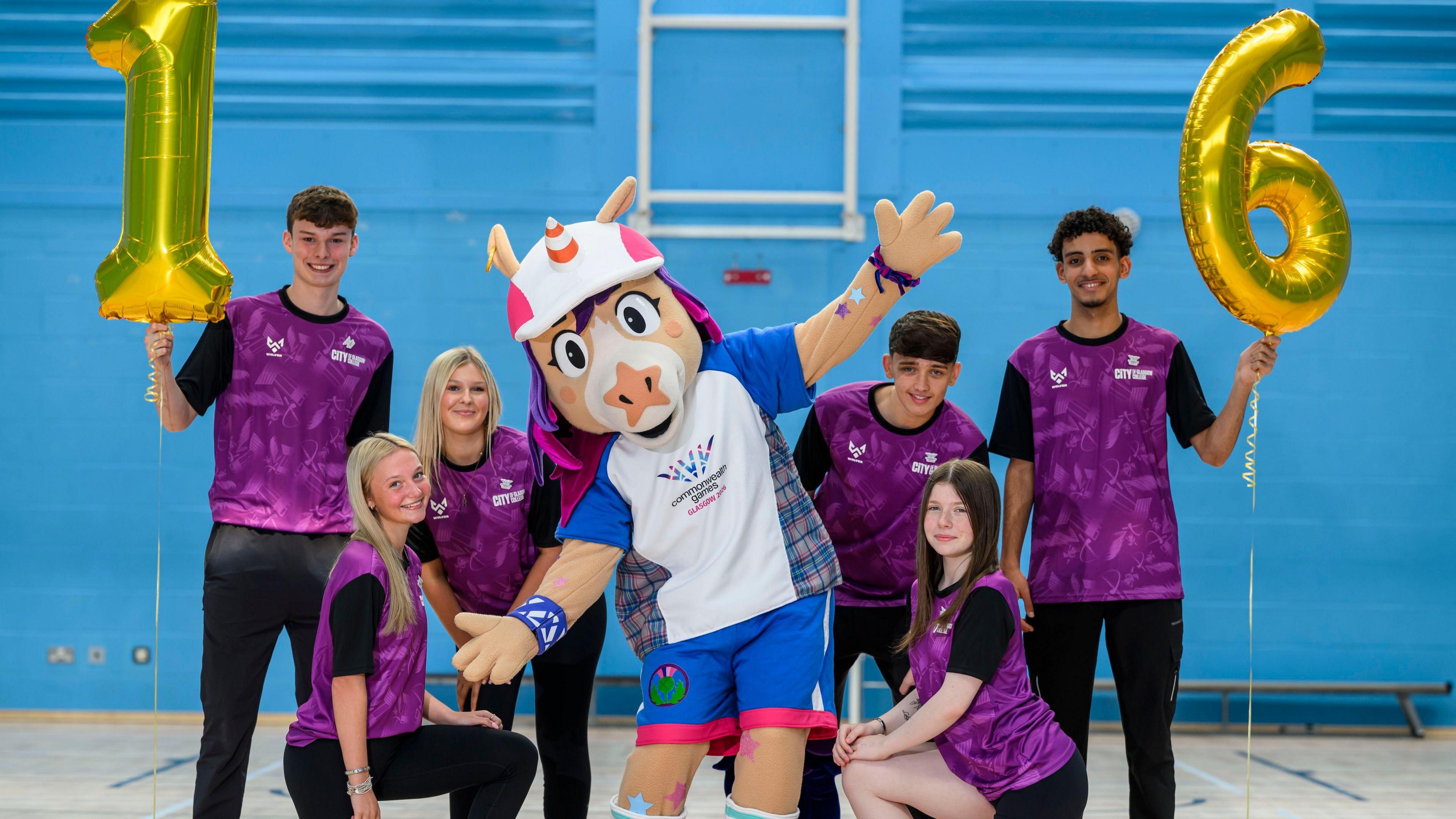 The Commonwealth Games mascot Finnie - a unicorn with a traffic cone for a horn and wearing a T-shirt and shorts saying Commonwealth Games on it, along with six volunteers for the Games - they are all wearing purple shirts and black trousers.