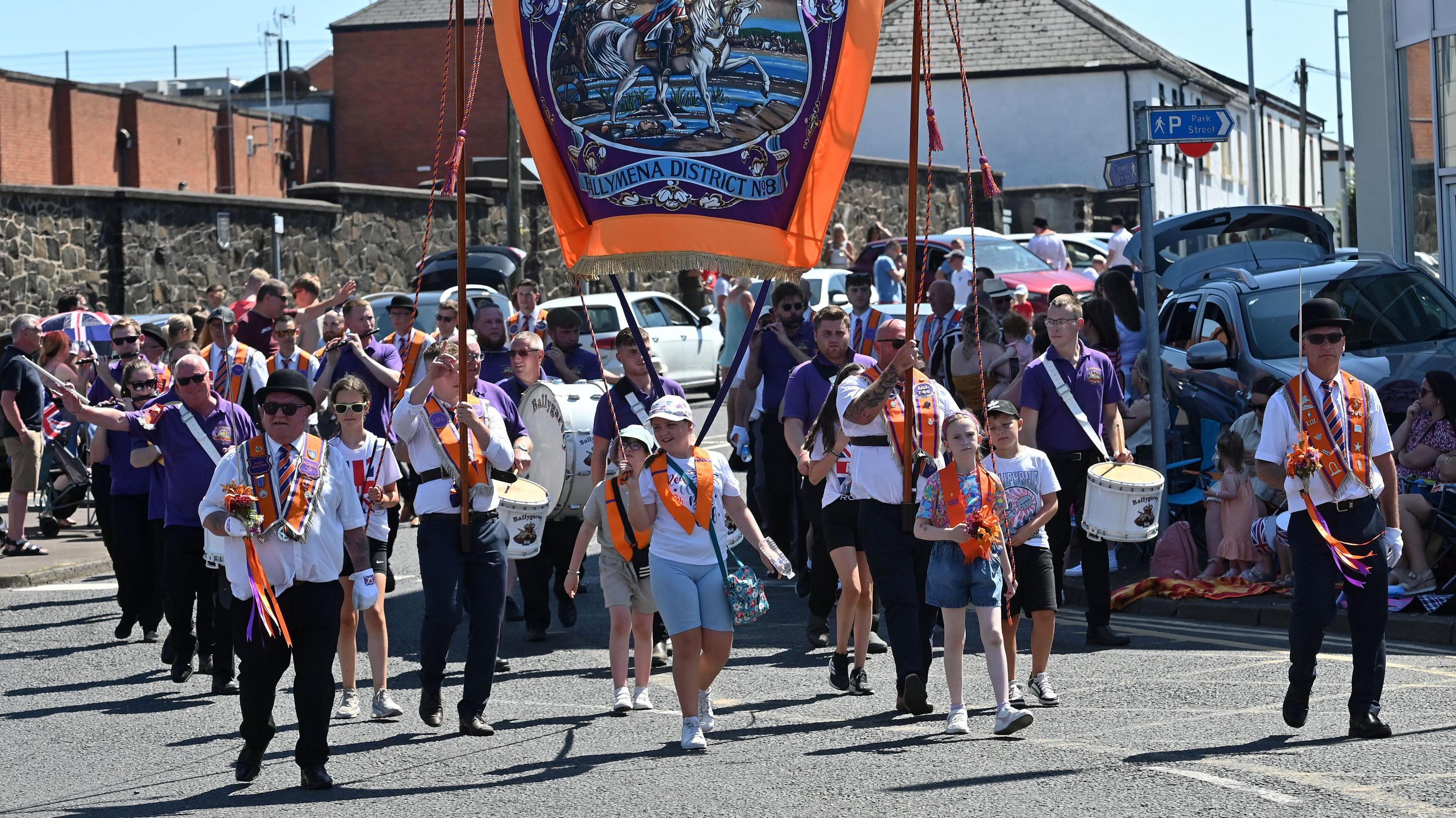 A group of people wearing orange sashes leading the parade, holding a large flag.