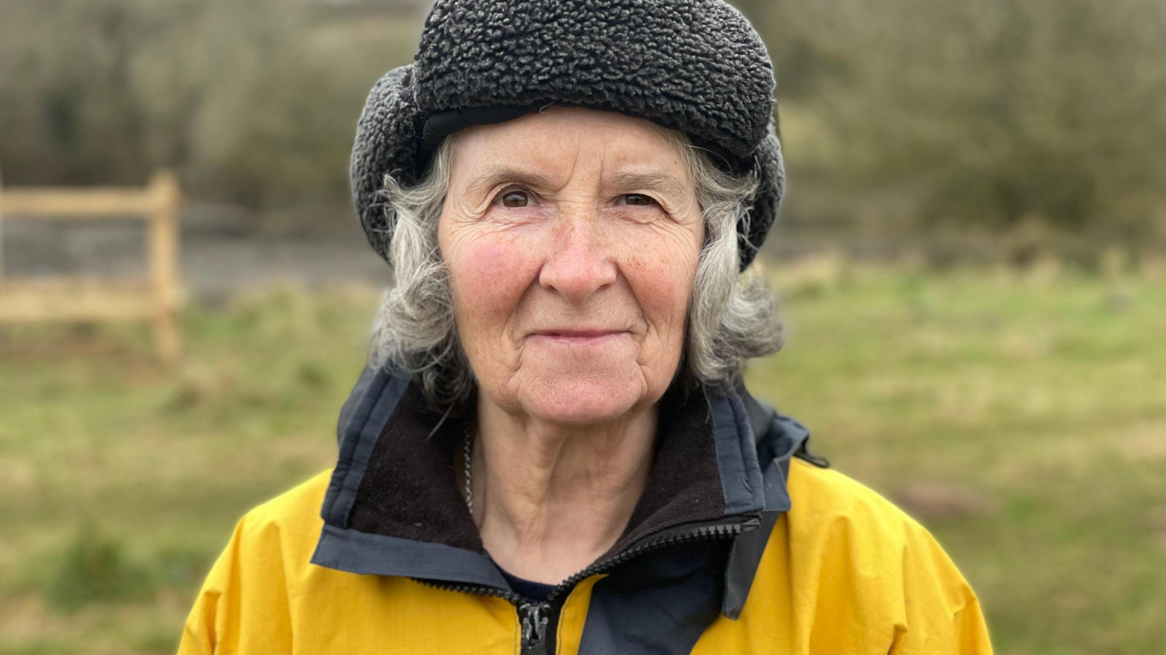 Gill looks at the camera while wearing a bright yellow rain coat and a fluffy grey hat with its edges folded up to reveal mid-length grey hair