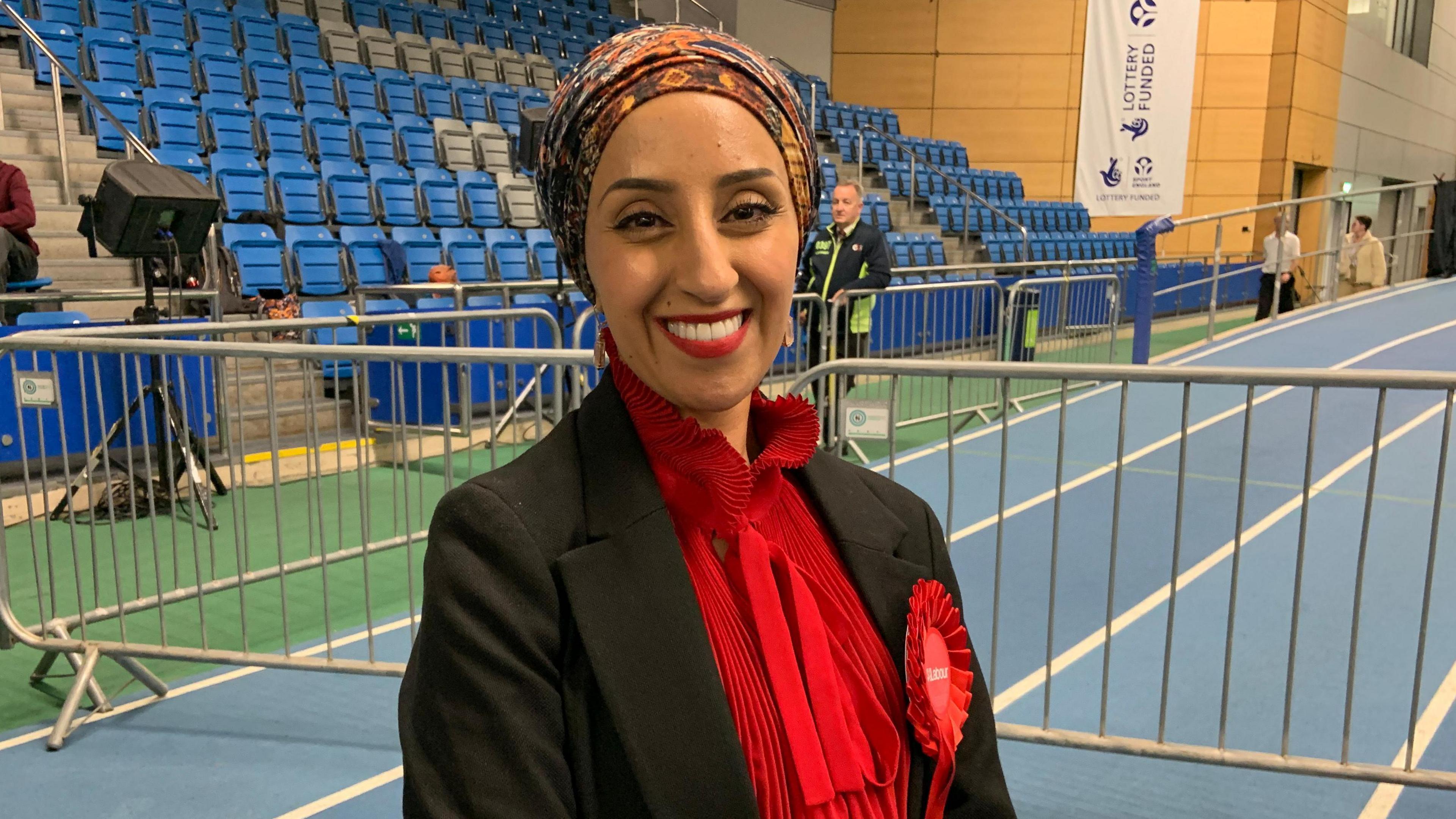 A woman wearing a coloured head scarf, black jacket and red shirt smiles into the camera. She is standing in a sports hall on a running track. Rows of blue seats are visible in the background.