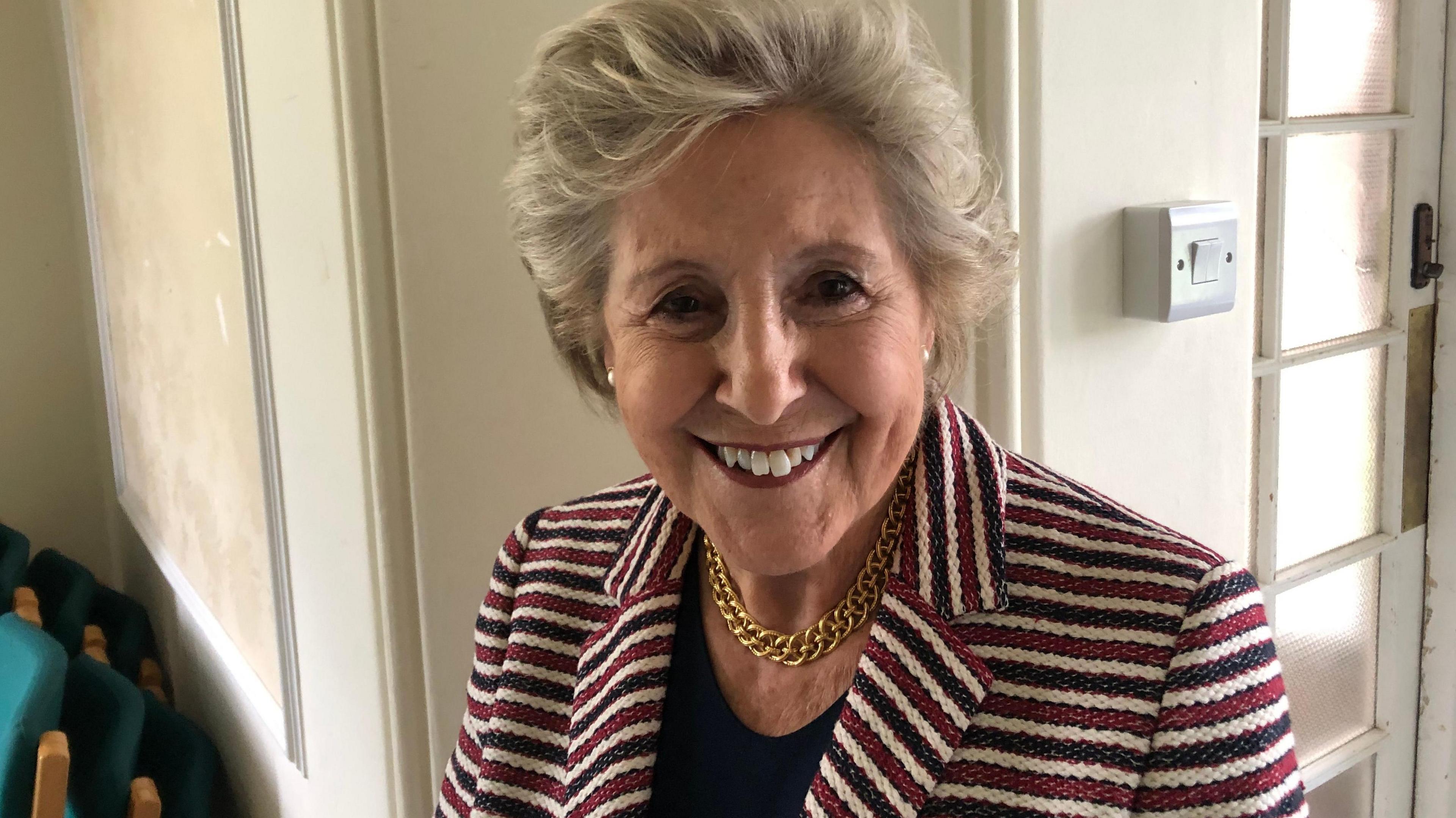 Dame Norma Major is smiling at the camera as she stands by a cream coloured wall. She is wearing a blazer with red, blue and white horizontal stripes and a black top underneath. She has light grey hair and also wears a gold chain link necklace, flat earrings.