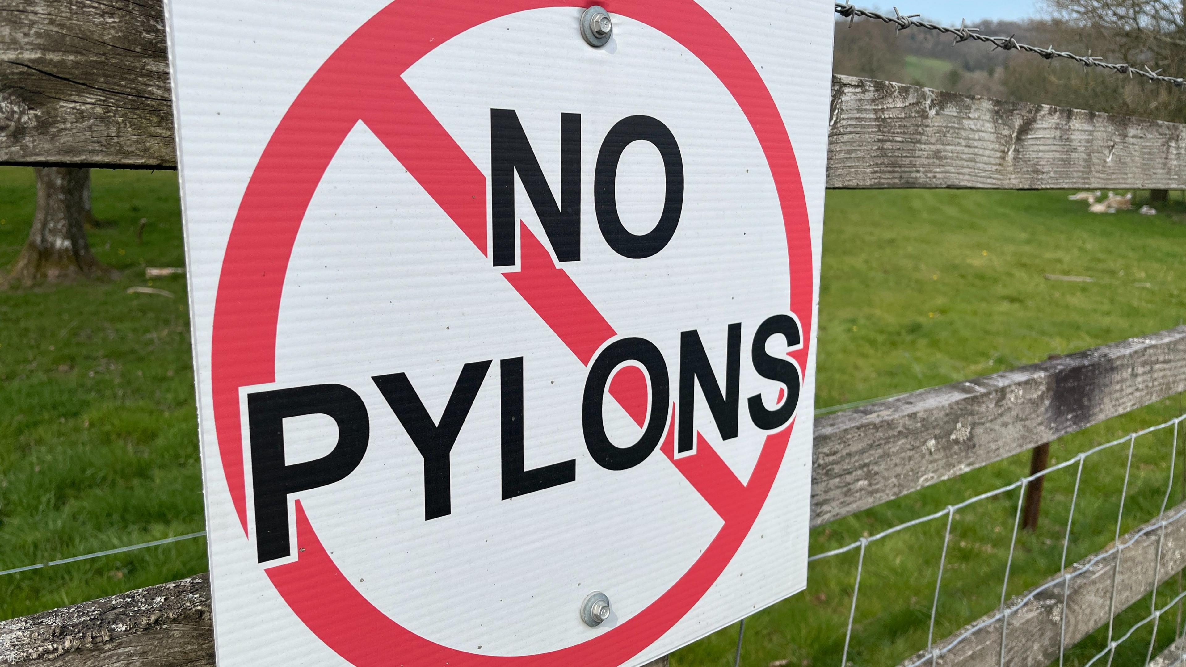 A sign that says NO PYLONS in front of a red circle with a line across it. The sign is attached to a wooden fence, a field is visible in the background.