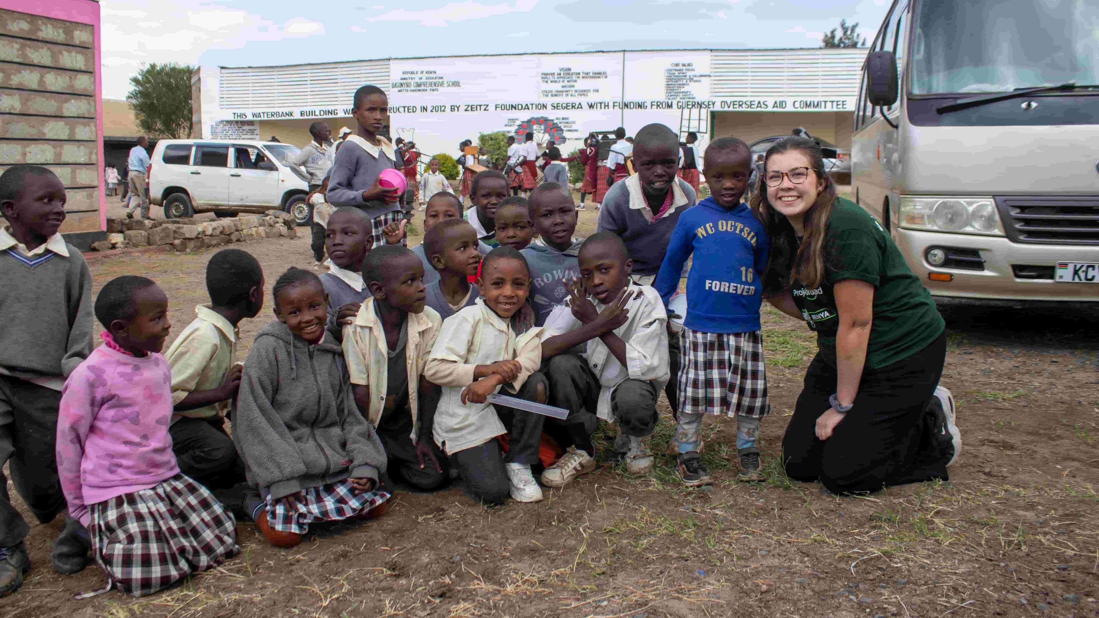 Ella McLaughlin (right) is smiling as she kneels on the ground alongside a dozen or so schoolchildren. She is wearing glasses and has her long hair tied back. Several of the youngsters are wearing grey jumpers or fleeces and white shirts. A van is in the background on the right-hand side.